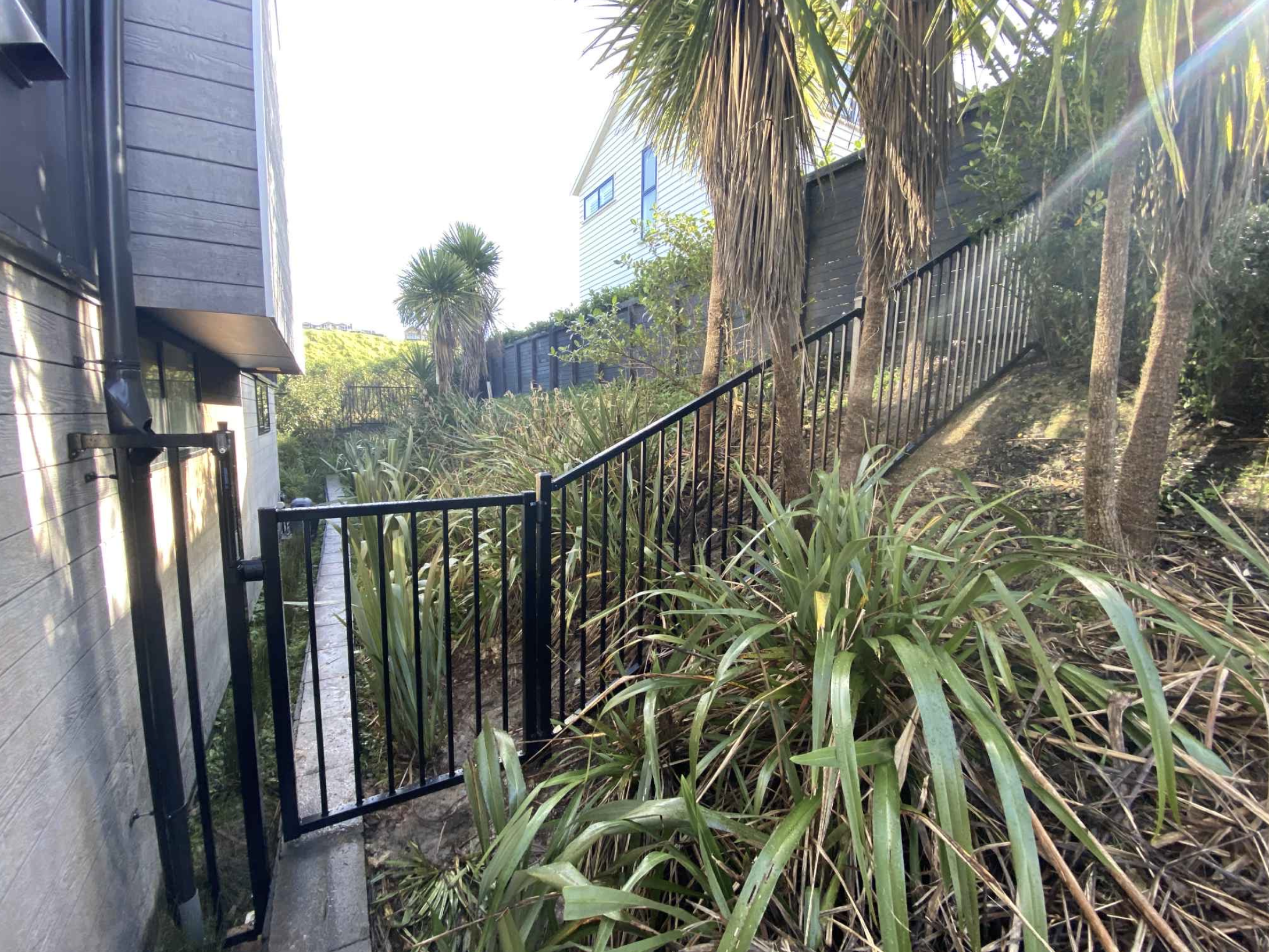 Side yard with black metal fence, lush green plants, and tall trees on a sloped terrain beside a gray house.