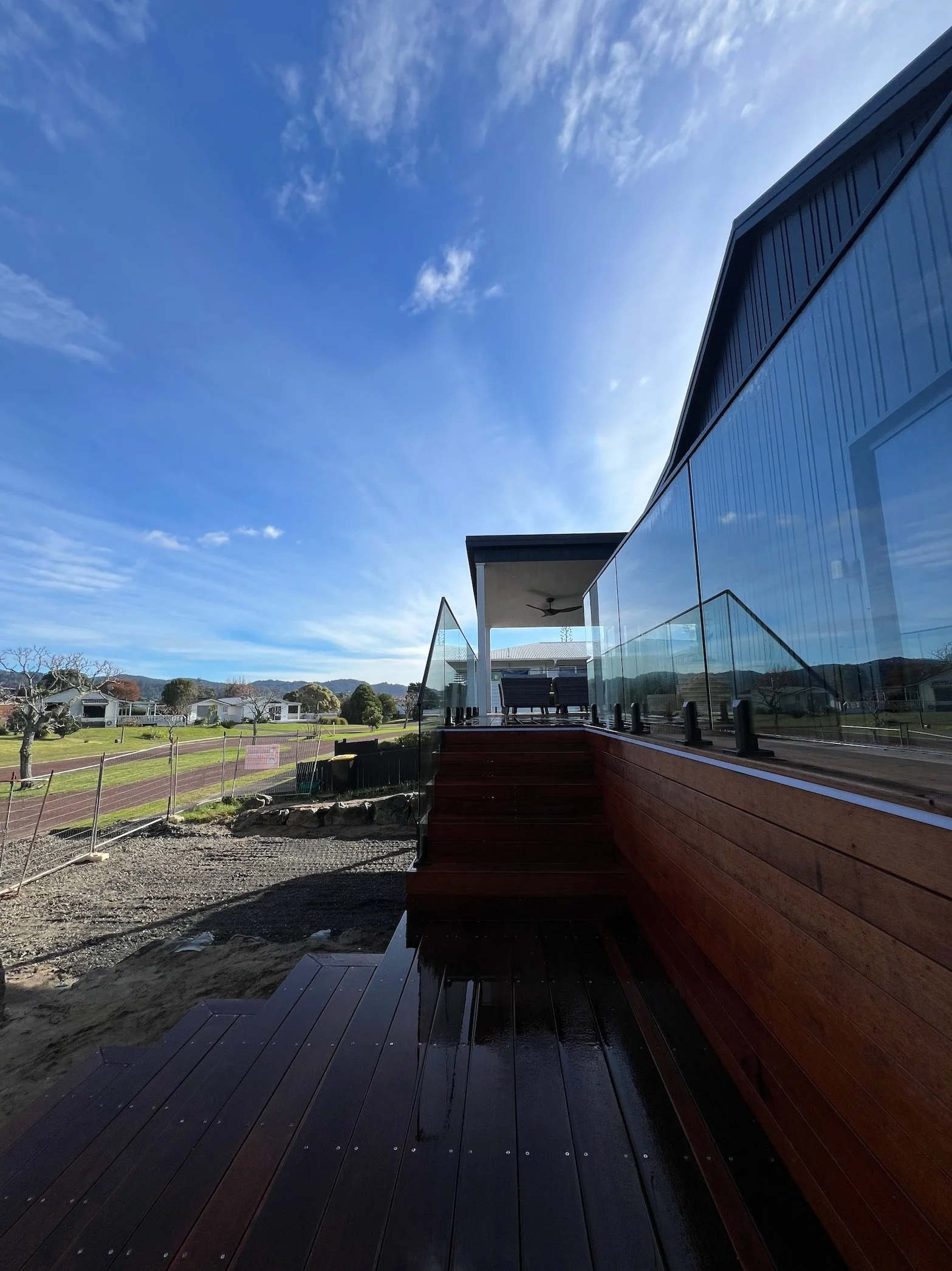 Modern house exterior with a wooden deck and glass railing, overlooking a landscape with a pathway, trees, and distant hills under a blue sky with scattered clouds.