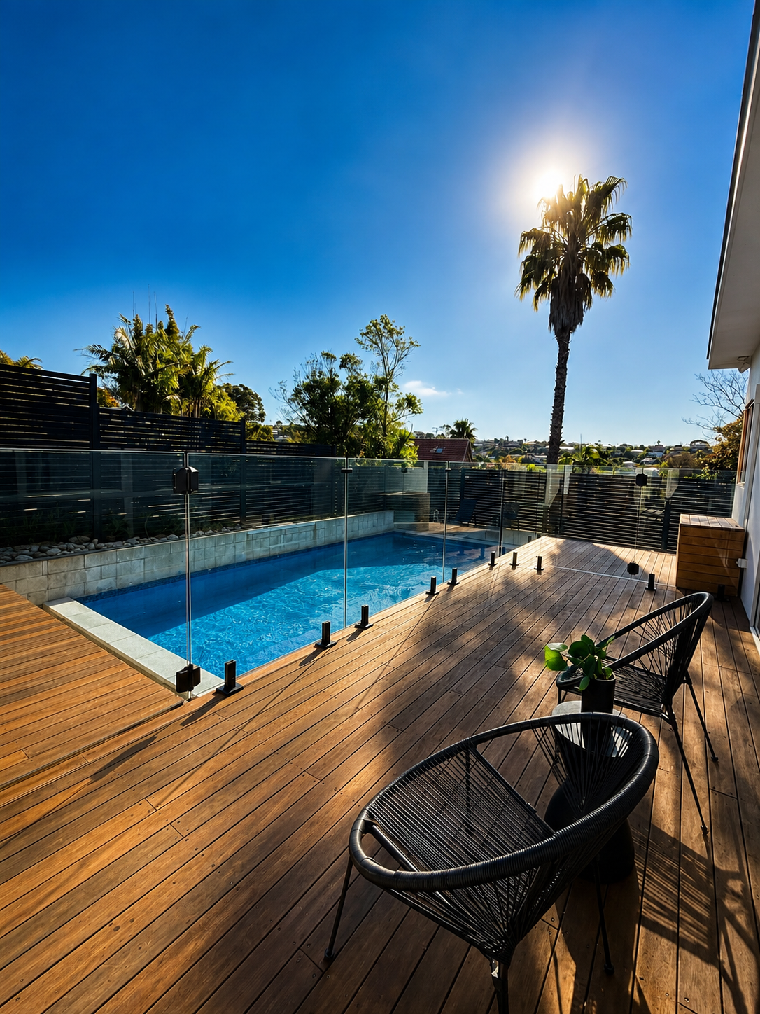 Backyard with a rectangular swimming pool, wooden deck, black outdoor chairs, and a potted plant, under a clear blue sky with the sun shining and palm trees in the background.