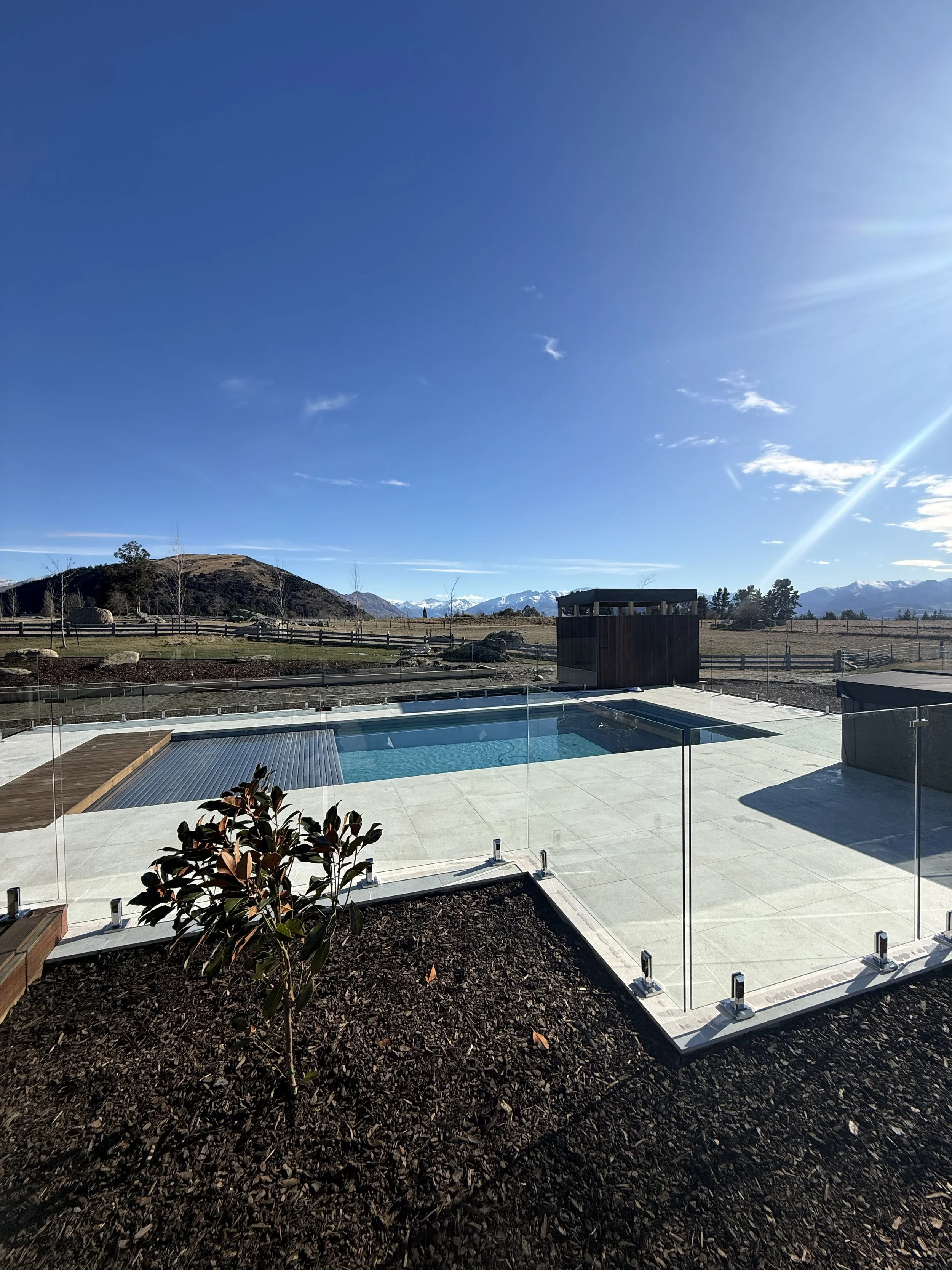 A backyard with a small swimming pool, surrounded by a concrete patio, a glass fence, and planted with mulch and plants. In the background, there are mountains and a clear blue sky with some clouds.