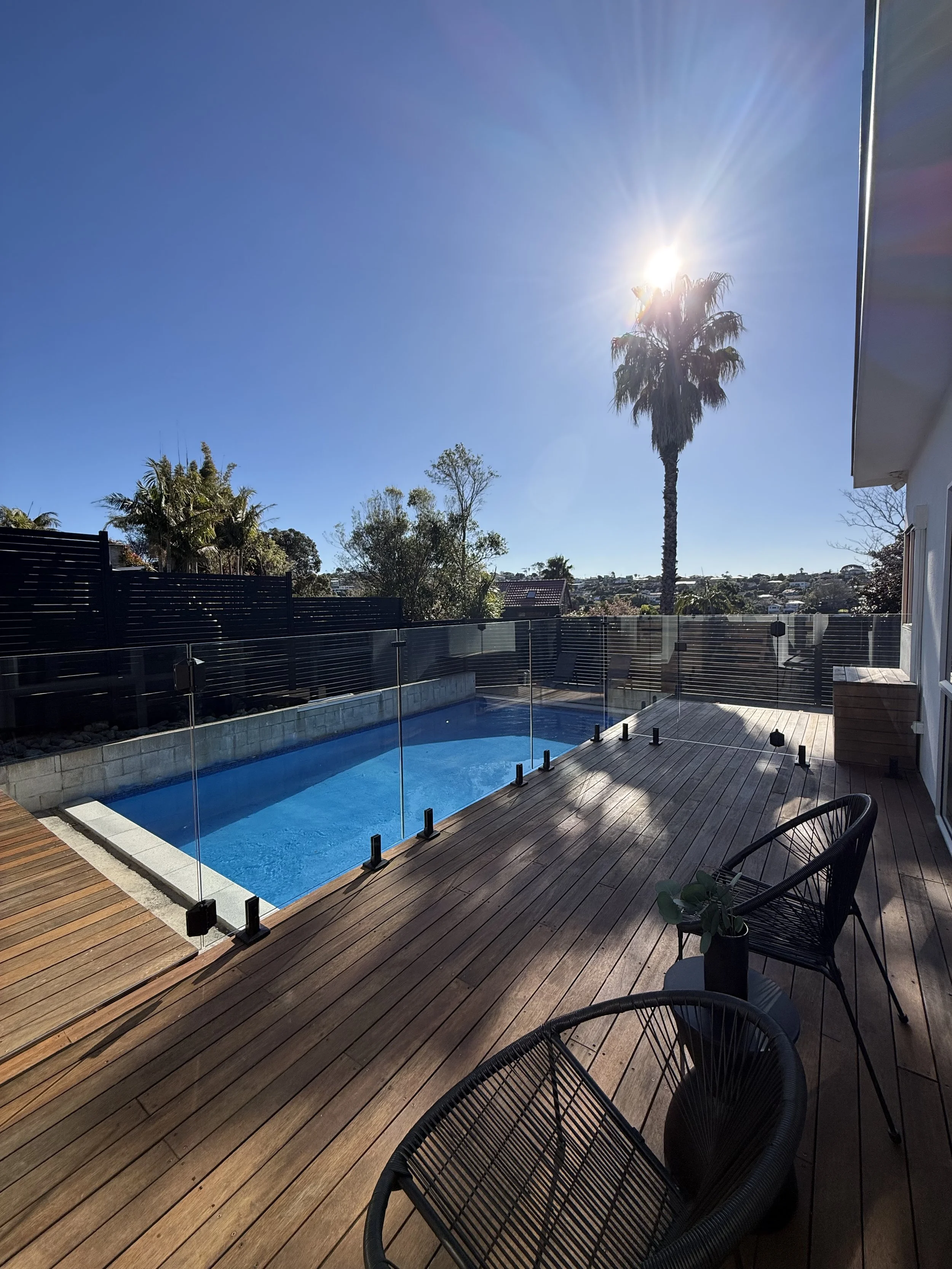 A backyard patio with a small swimming pool, wooden deck, black fence, patio chairs, and a palm tree with the sun shining brightly in the clear blue sky.