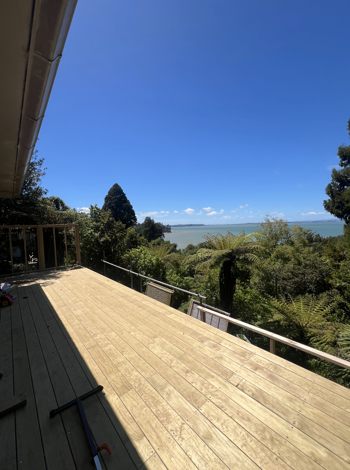 View from a wooden deck overlooking lush green trees, with calm water and a blue sky in the background.