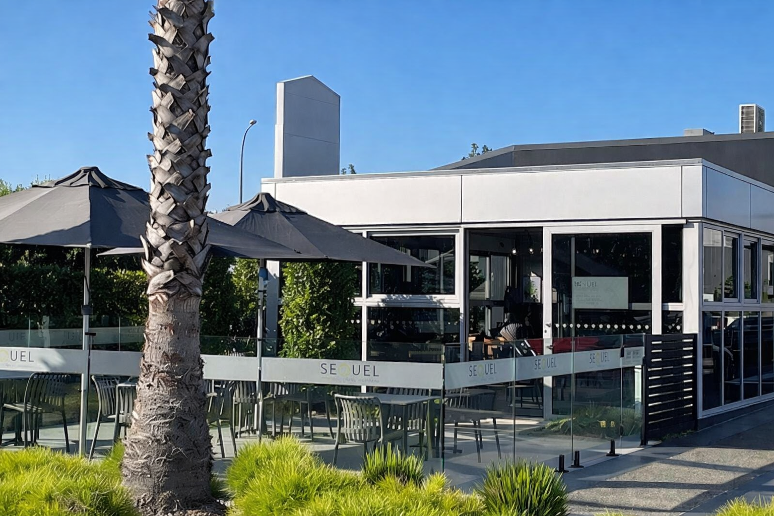 Exterior view of a modern restaurant called SEULE with outdoor seating, large umbrellas, and a palm tree in the foreground, under a clear blue sky.