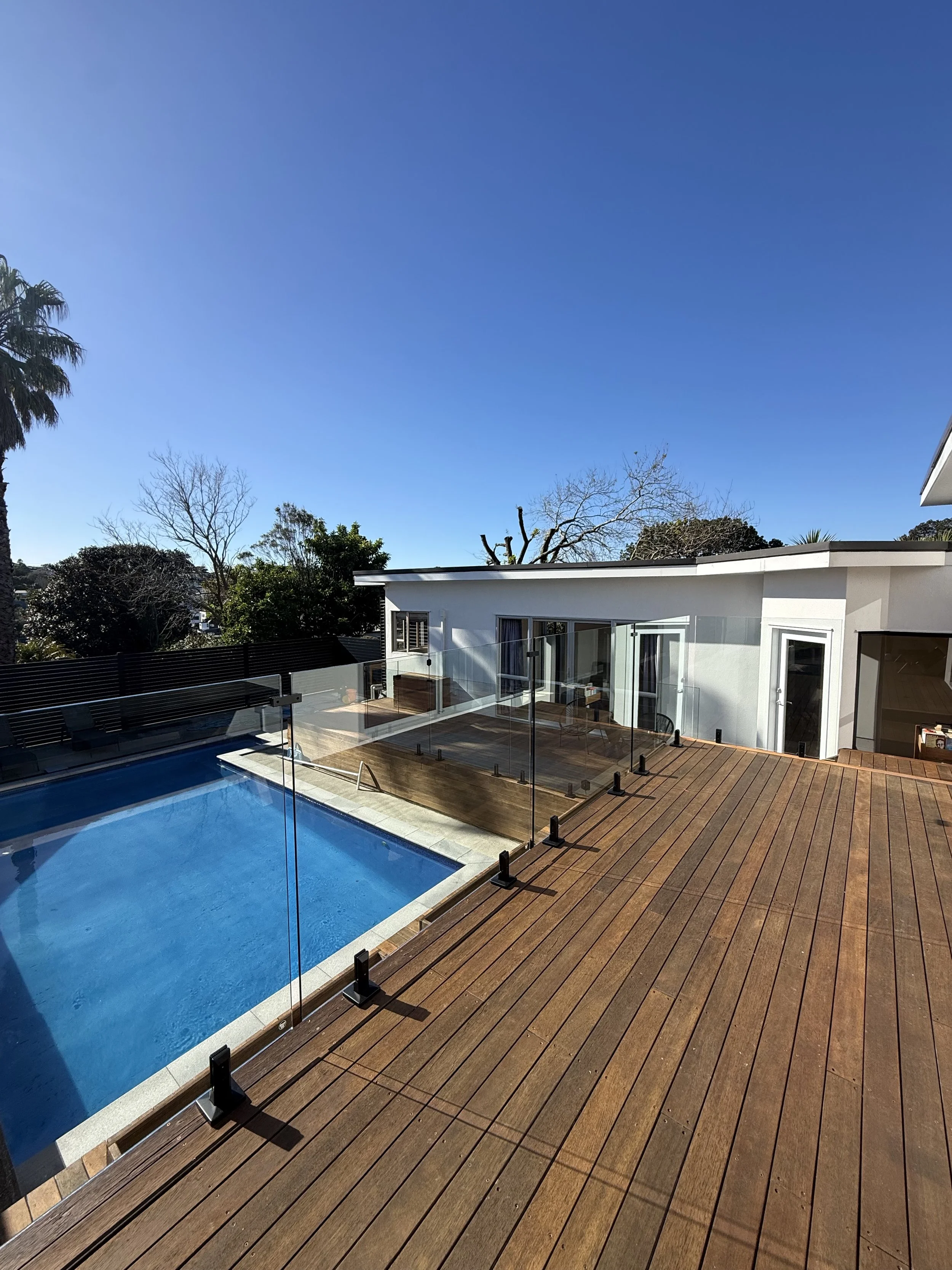 A modern house with large windows and a spacious wooden deck area, next to a swimming pool with a glass fence, under a clear blue sky with some trees in the background.