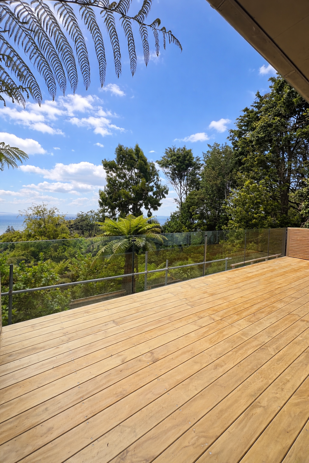 A wooden deck with glass railing overlooking lush green trees and a blue sky with scattered clouds.