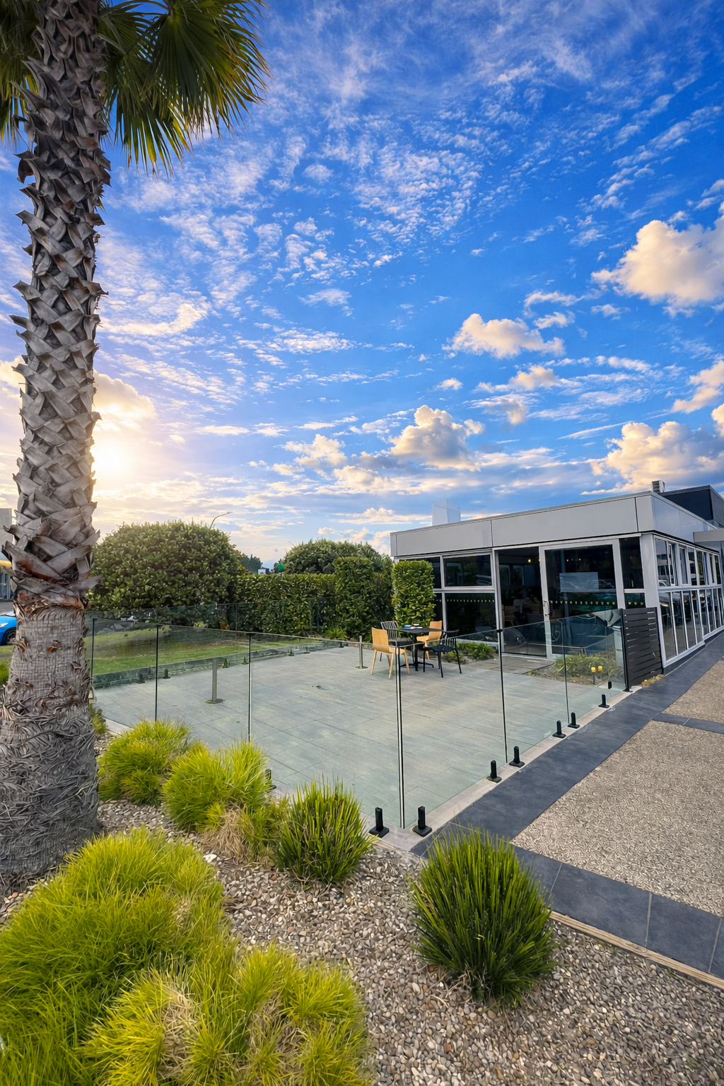 A modern building with large glass windows, an outdoor patio with table and chairs, surrounded by landscaping including palm trees, bushes, and drought-tolerant plants. The sky is blue with scattered clouds, suggesting late afternoon or early evening