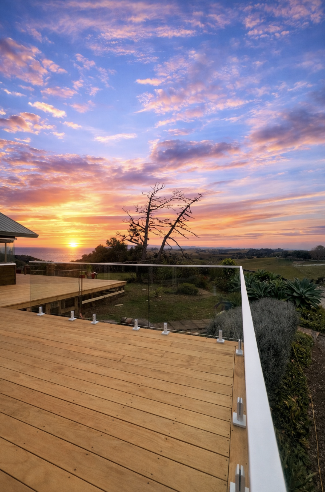 A sunset view from a wooden deck with glass railing, overlooking a green landscape with a barren tree, a few bushes, and a distant ocean or large body of water under a colorful sky with pink, purple, and orange clouds.