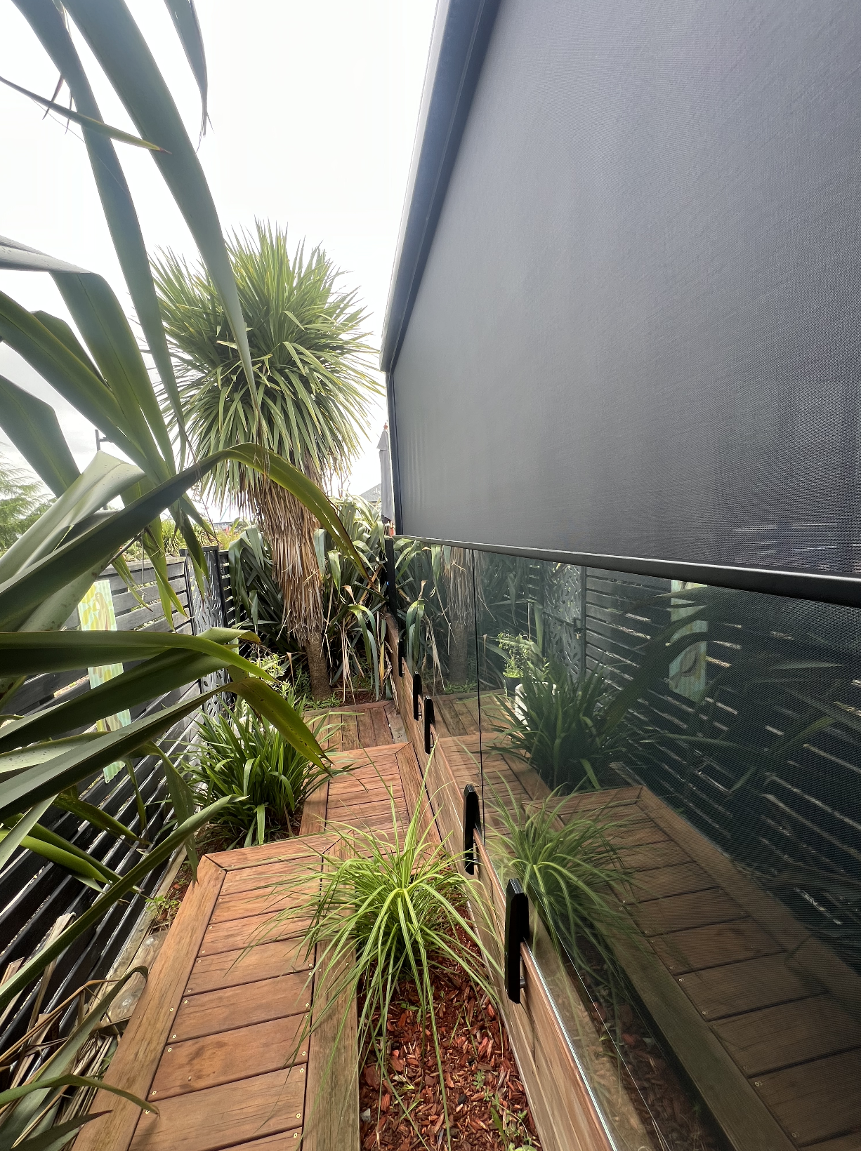 Small outdoor balcony with wooden flooring, green potted plants, and a large screen or shade covering part of the balcony, surrounded by trees and plants.
