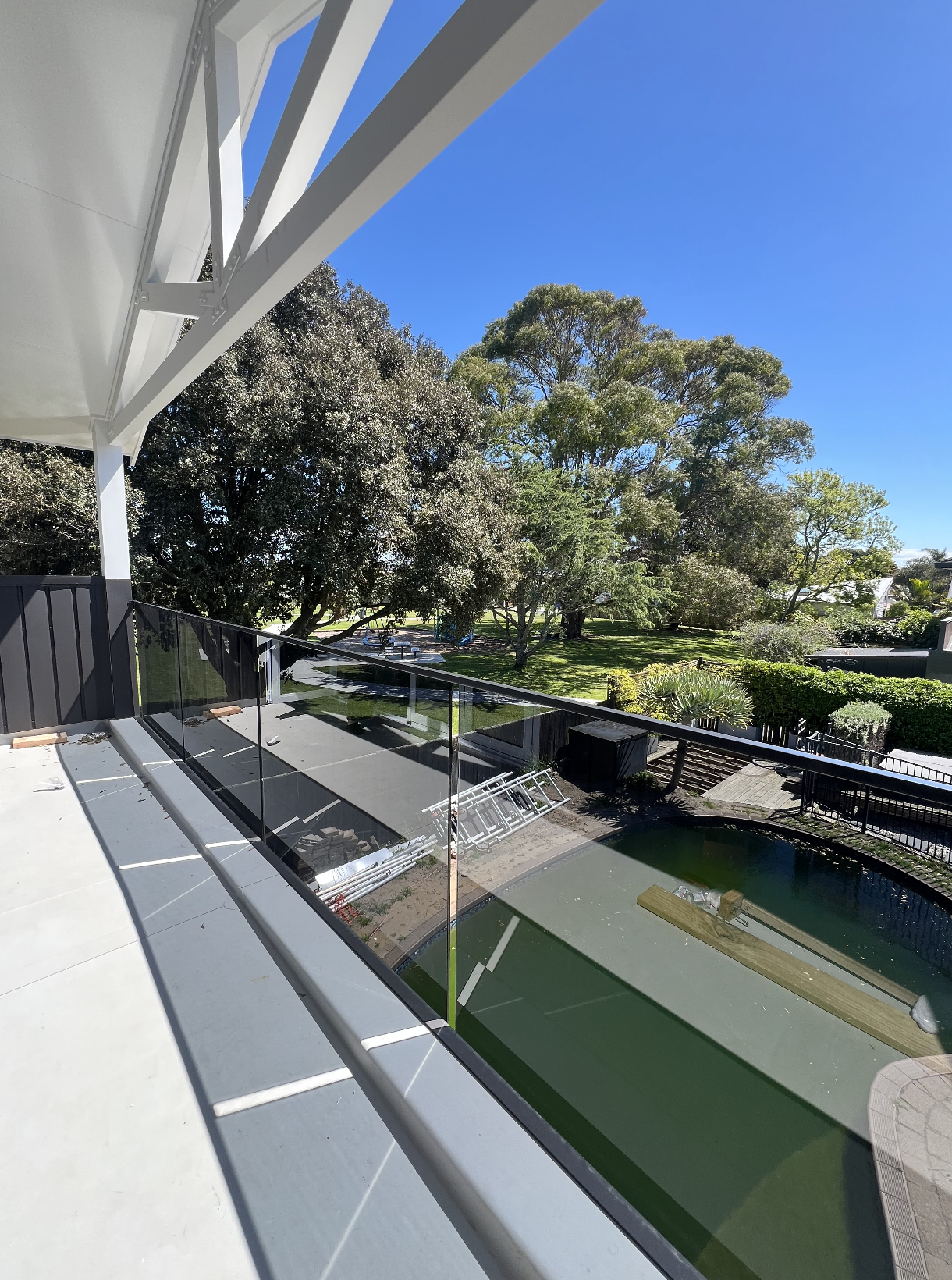 View from a balcony showing a green backyard with trees, a small pool, and construction materials on the ground outside.