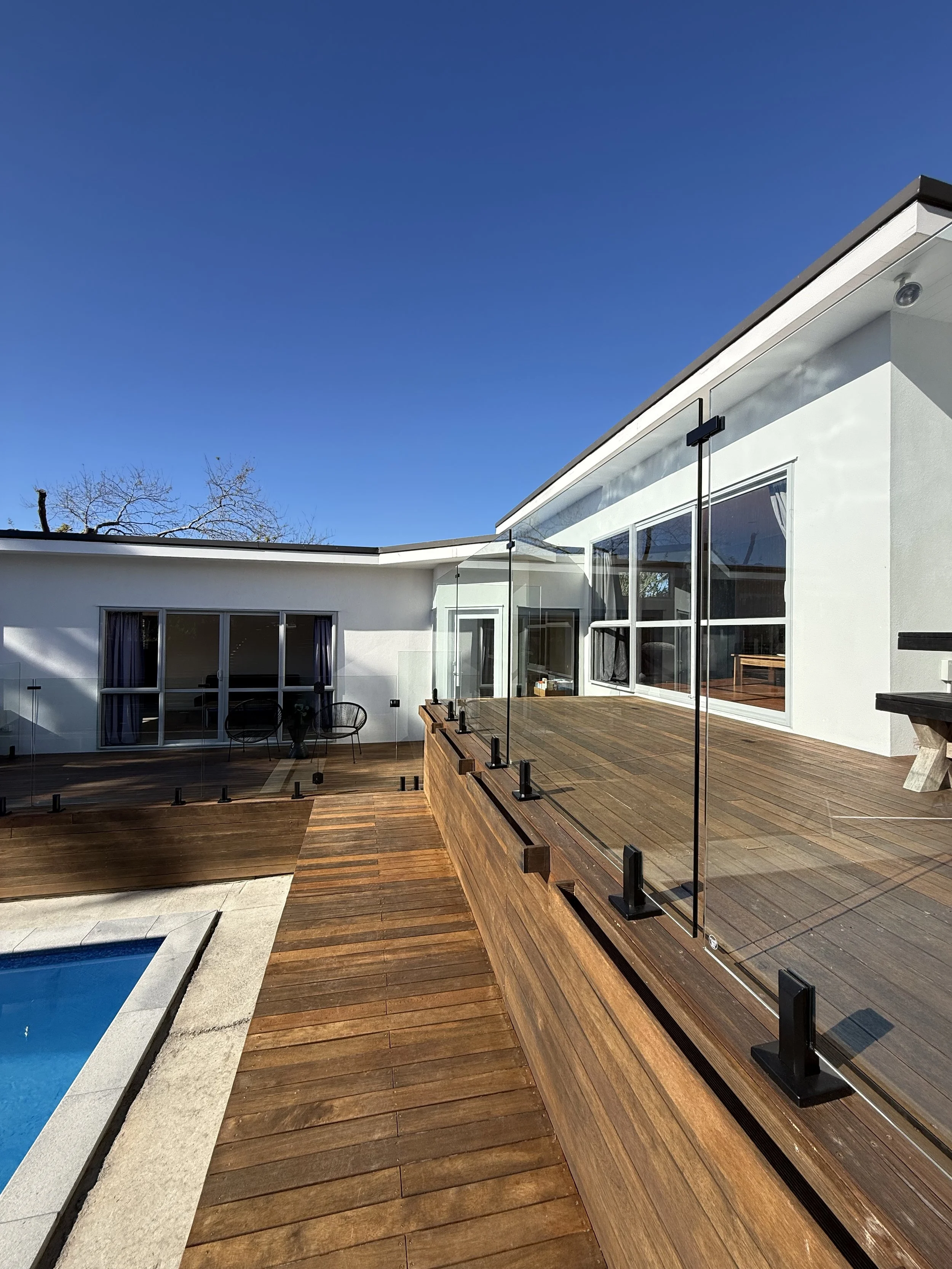 Modern house with wooden deck, glass railing, sliding glass doors, and swimming pool in the yard under a clear blue sky.