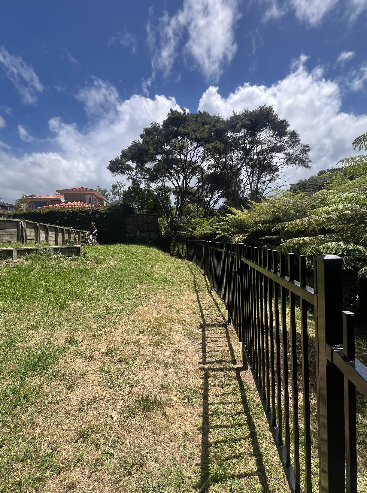 A grassy pathway bordered by a black metal fence on the right, lush green trees on the right, and a person walking in the distance on the left, under a partly cloudy blue sky.