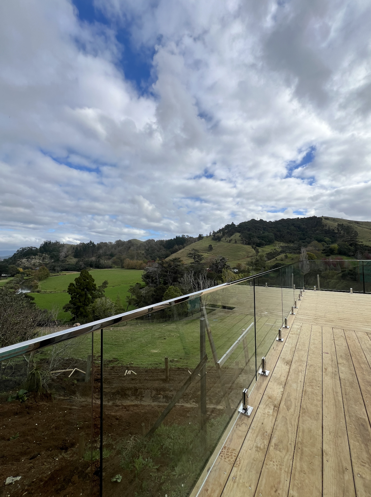 View from a wooden deck with glass railing overlooking green rolling hills and cloudy sky.