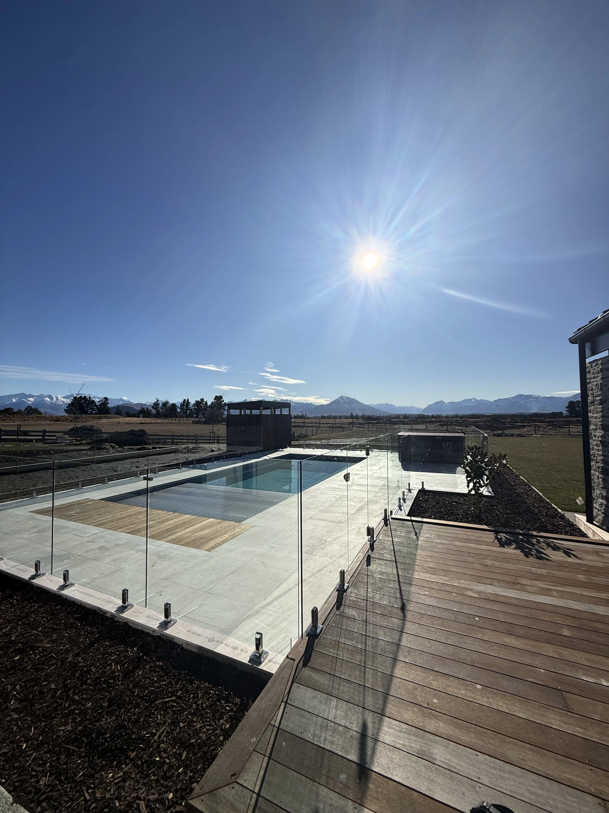 An outdoor scene showing a backyard with a swimming pool, wooden deck, and a glass fence, with mountain ranges in the distance under a bright sun in a clear blue sky.