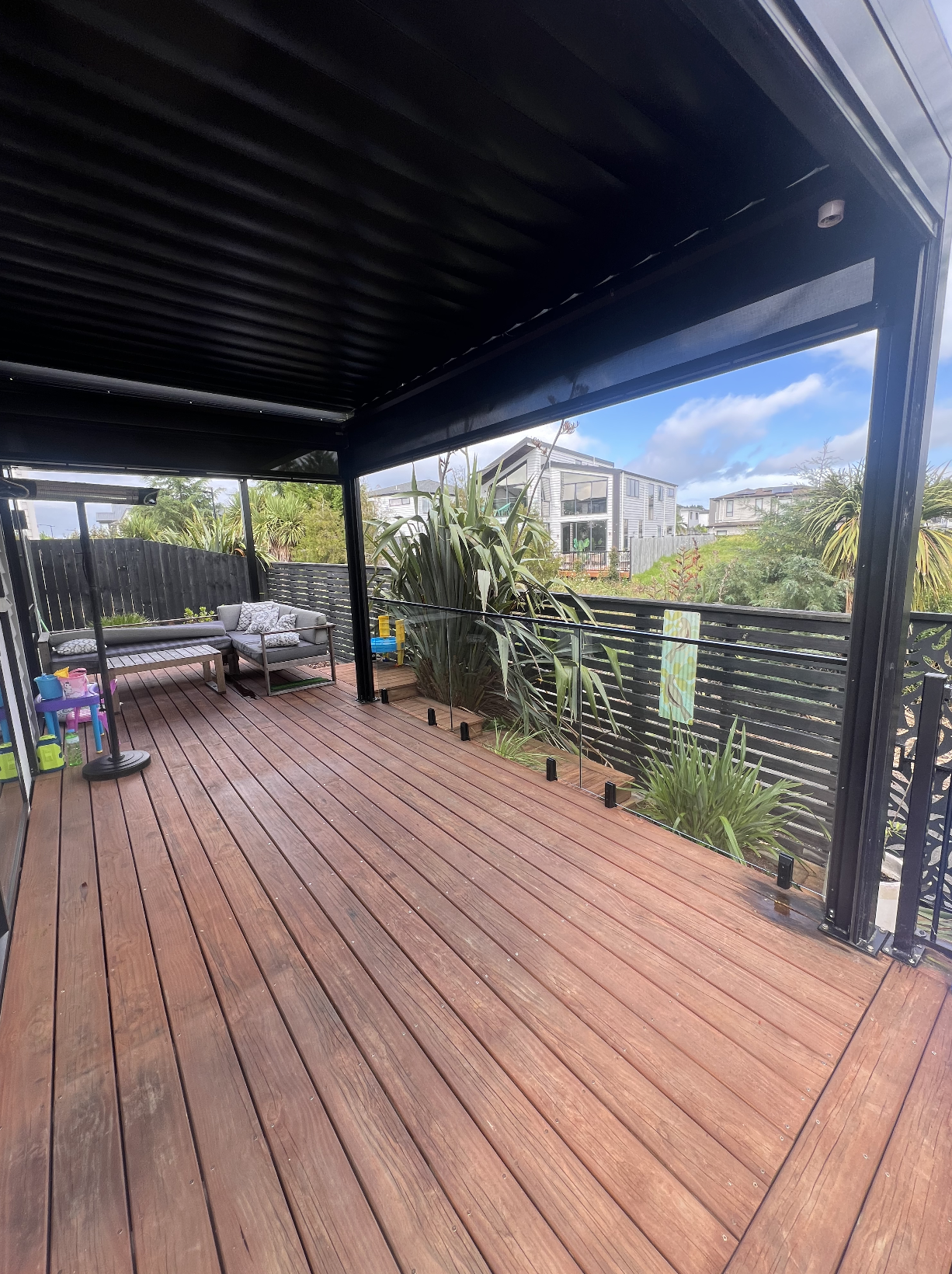 Empty wooden deck with outdoor seating, plants, and a black metal railing under a black metal roof, with a view of houses and trees in the background.