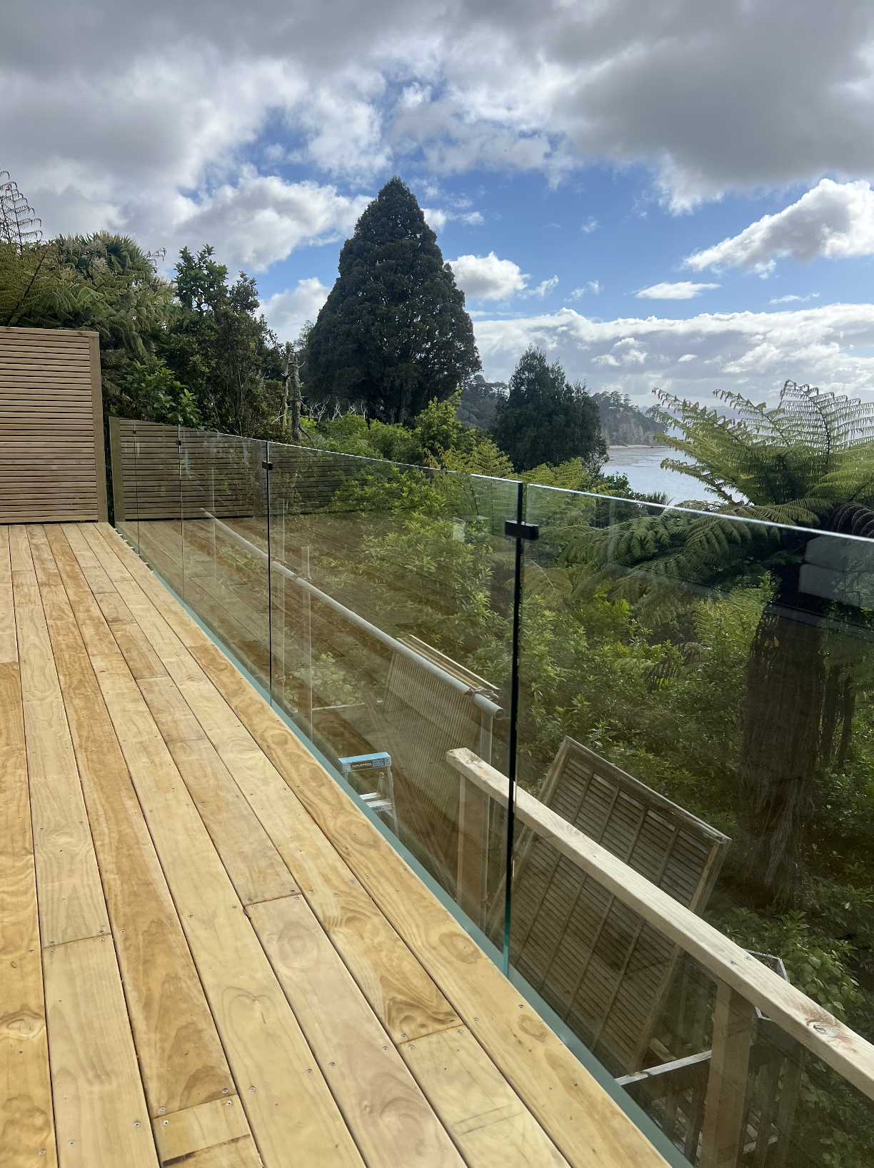 A wooden deck with a glass railing overlooking a lush green landscape with trees and bushes, under a partly cloudy sky.