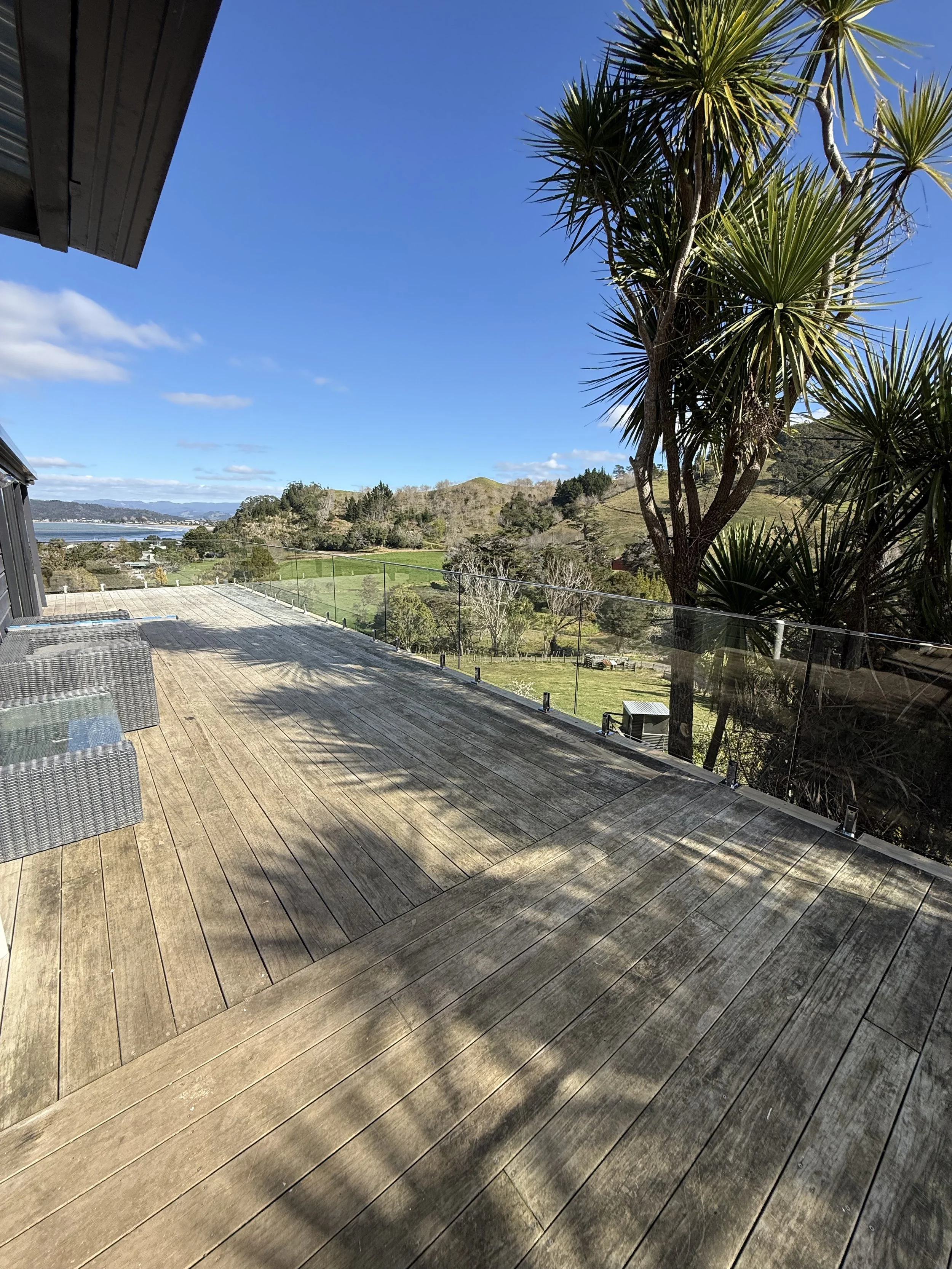 View of a wooden deck with outdoor furniture, palm trees, rolling hills, and a bright blue sky with some clouds.