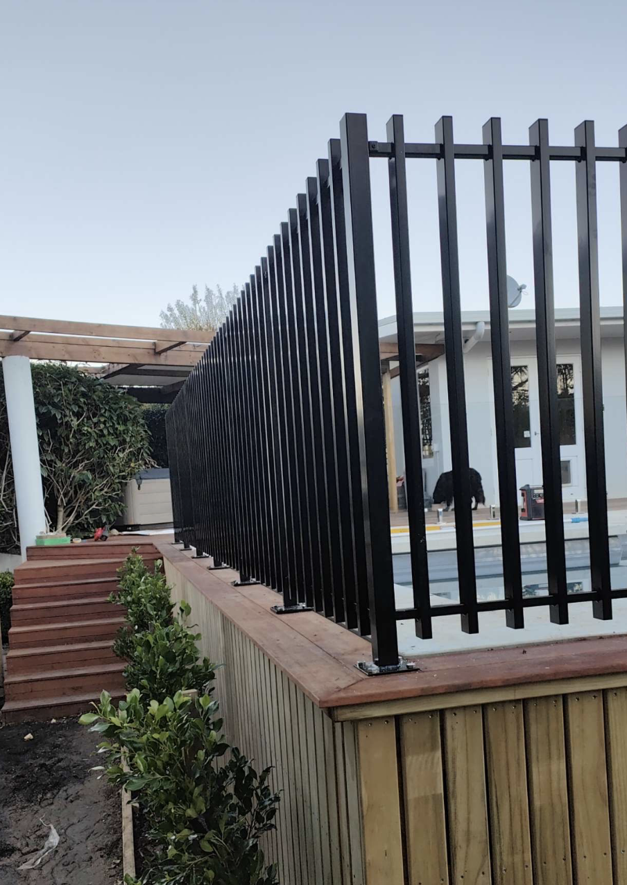 View of a black metal fence installed around a backyard patio with wooden steps leading up to the area, and some plants in the foreground.