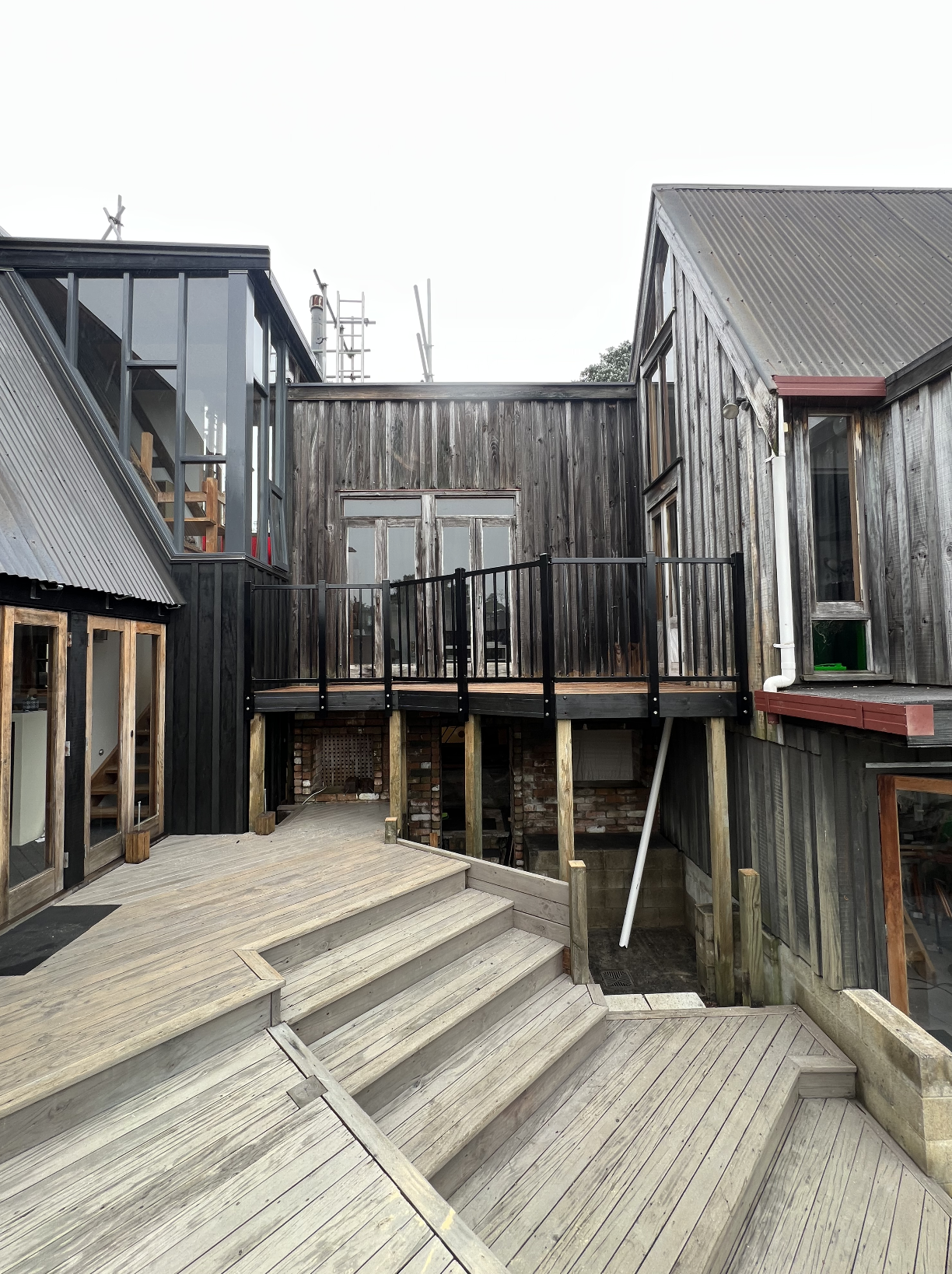 Backyard with stairs leading to an elevated deck, surrounded by wooden houses with a mix of weathered and newer wood siding, some large glass windows, and a cloudy sky.