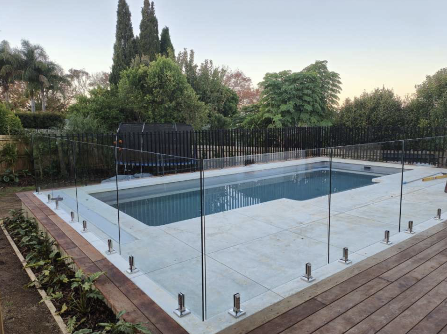 A backyard with a rectangular swimming pool with a glass fence around it, surrounded by a wooden deck and lush green trees and plants in the background during late afternoon or early evening.