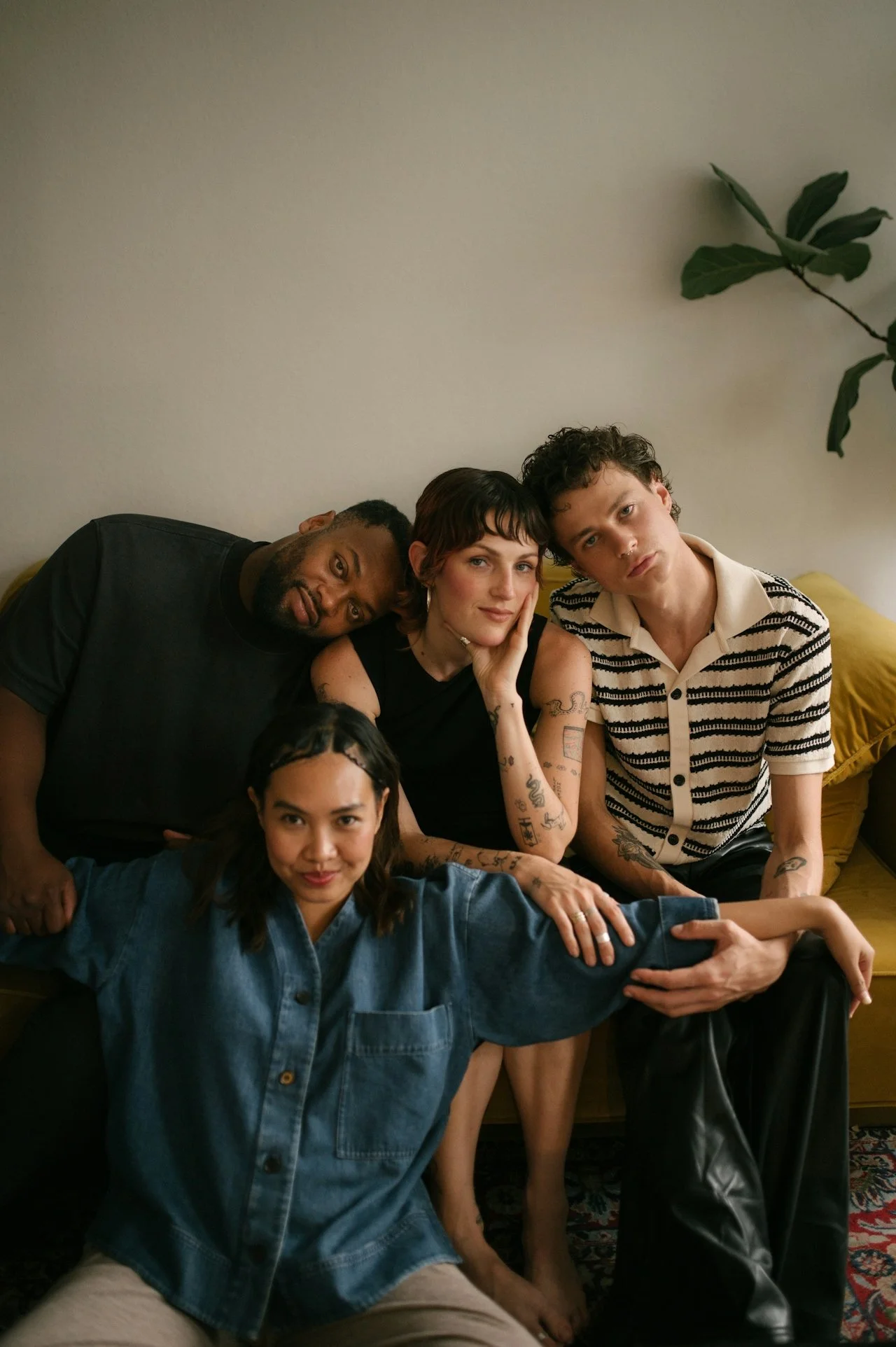 A group of five diverse young adults sitting on a yellow couch against a plain wall, with a large green plant in the background, posing casually for a photo.
