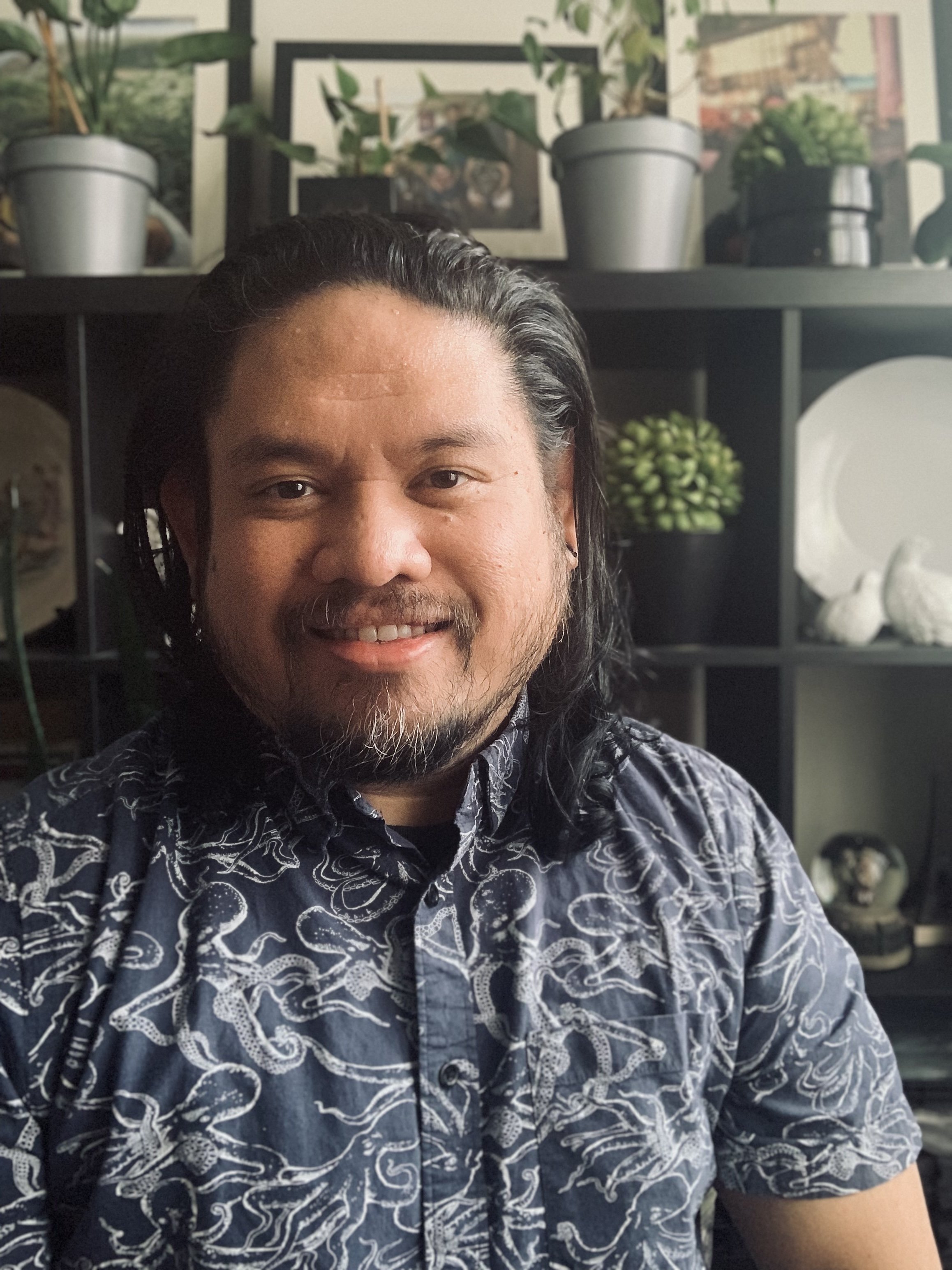 A man with long black hair, a beard, and a mustache smiling at the camera, wearing a patterned short-sleeve shirt, with a black shelf and various decorative items and plants in the background.