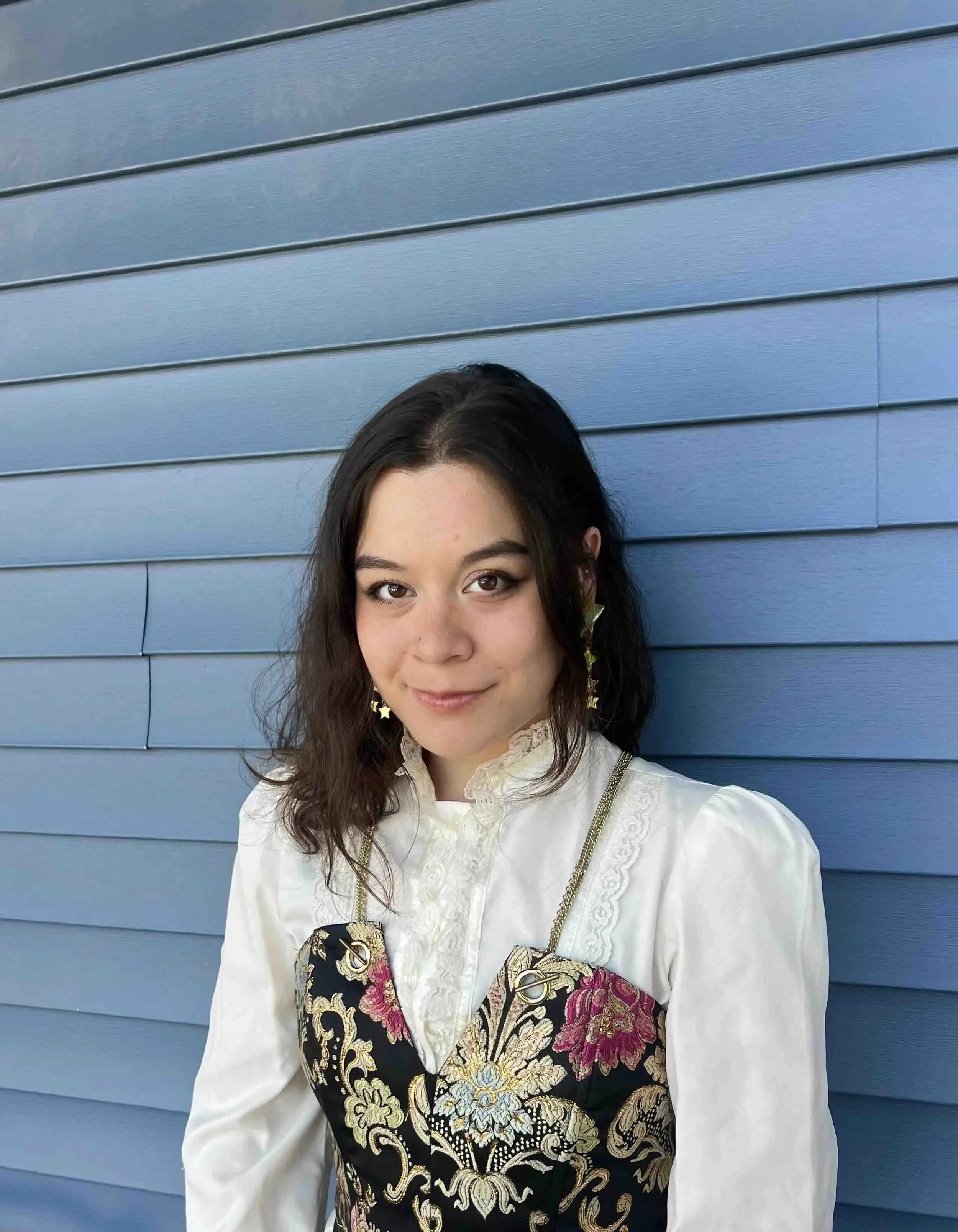 Rhys Hare, a young woman with shoulder-length dark hair, wearing a white blouse with ruffled collar, decorative gold earrings, and a black embroidered dress with floral designs, standing against a blue wooden wall.