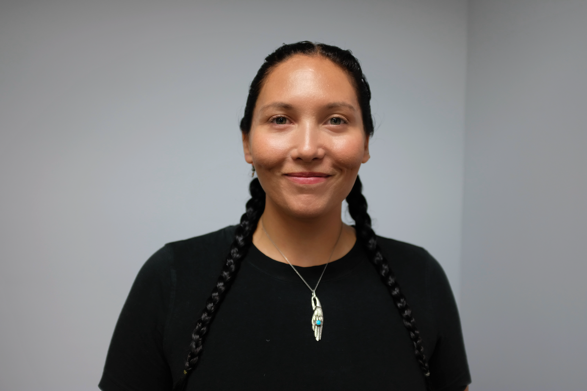 Laura Witkop, a woman with dark hair in braids, wearing a black shirt and a silver necklace with a feather-shaped pendant, smiling at the camera against a plain neutral background.
