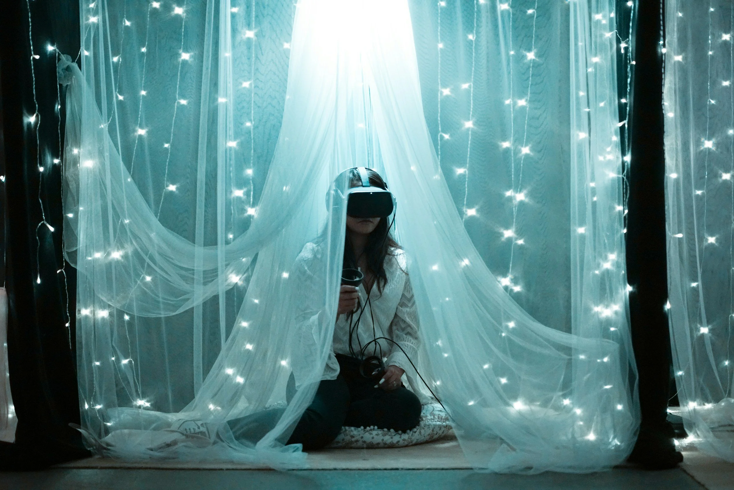 Young woman sitting on a cushion wearing VR goggles and draped in a cozy starlit tulle curtain