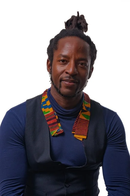 Quincy Adams, a man with dreadlocks tied up, wearing a blue long sleeve shirt, a dark vest, and a colorful kente cloth collar.