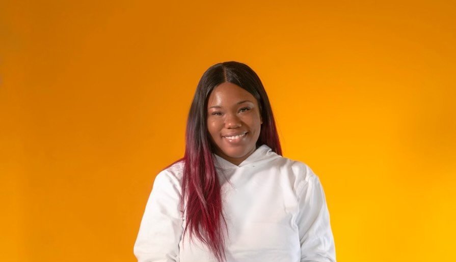 A young woman with long black hair with pink tips, wearing a white hoodie, smiling against a plain orange background.
