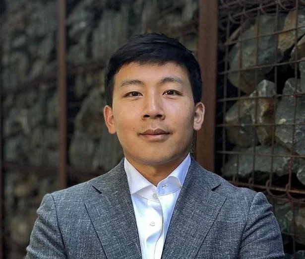 Huezong Yang, a smiling man in a grey suit with a white shirt standing outdoors against a background of rocks and metal fencing.