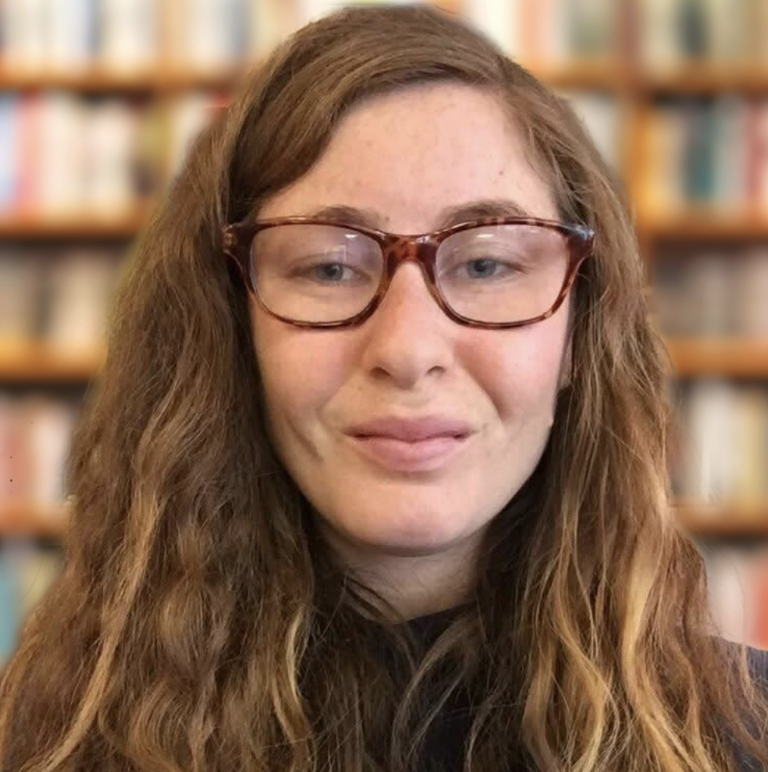 Shannon Hassett, a woman with long wavy brown hair and glasses, standing in front of a bookshelf.