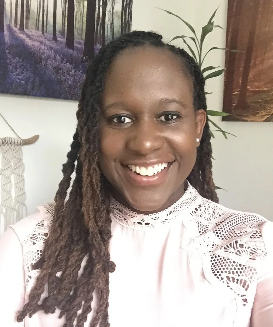 Shari Appollon, a smiling woman with dreadlocks wearing a light pink top with lace details, standing indoors with framed pictures of a forest and plants in the background.