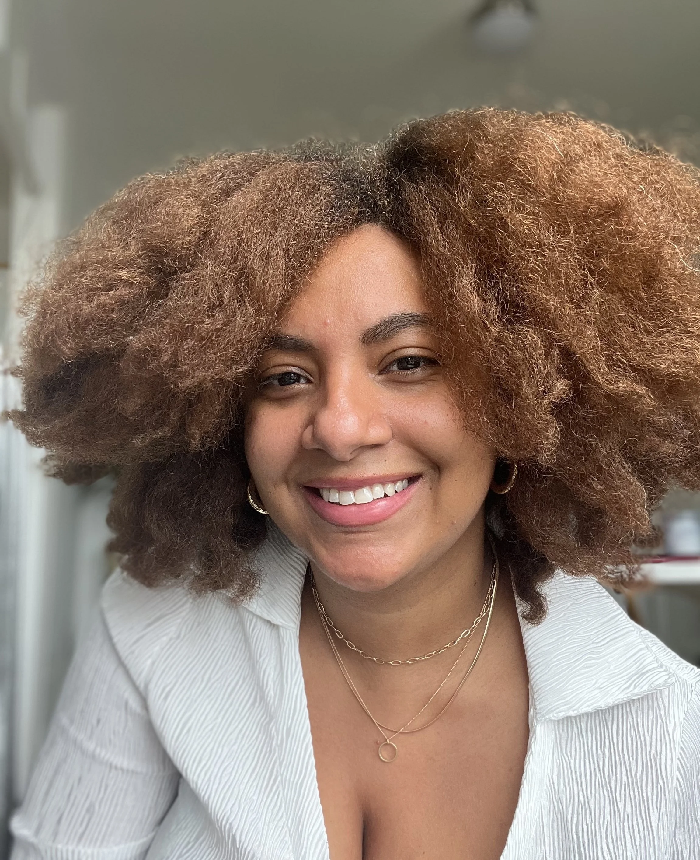 A woman with voluminous curly brown hair smiling, wearing a white top with a deep neckline and layered gold necklaces, in an indoor setting.