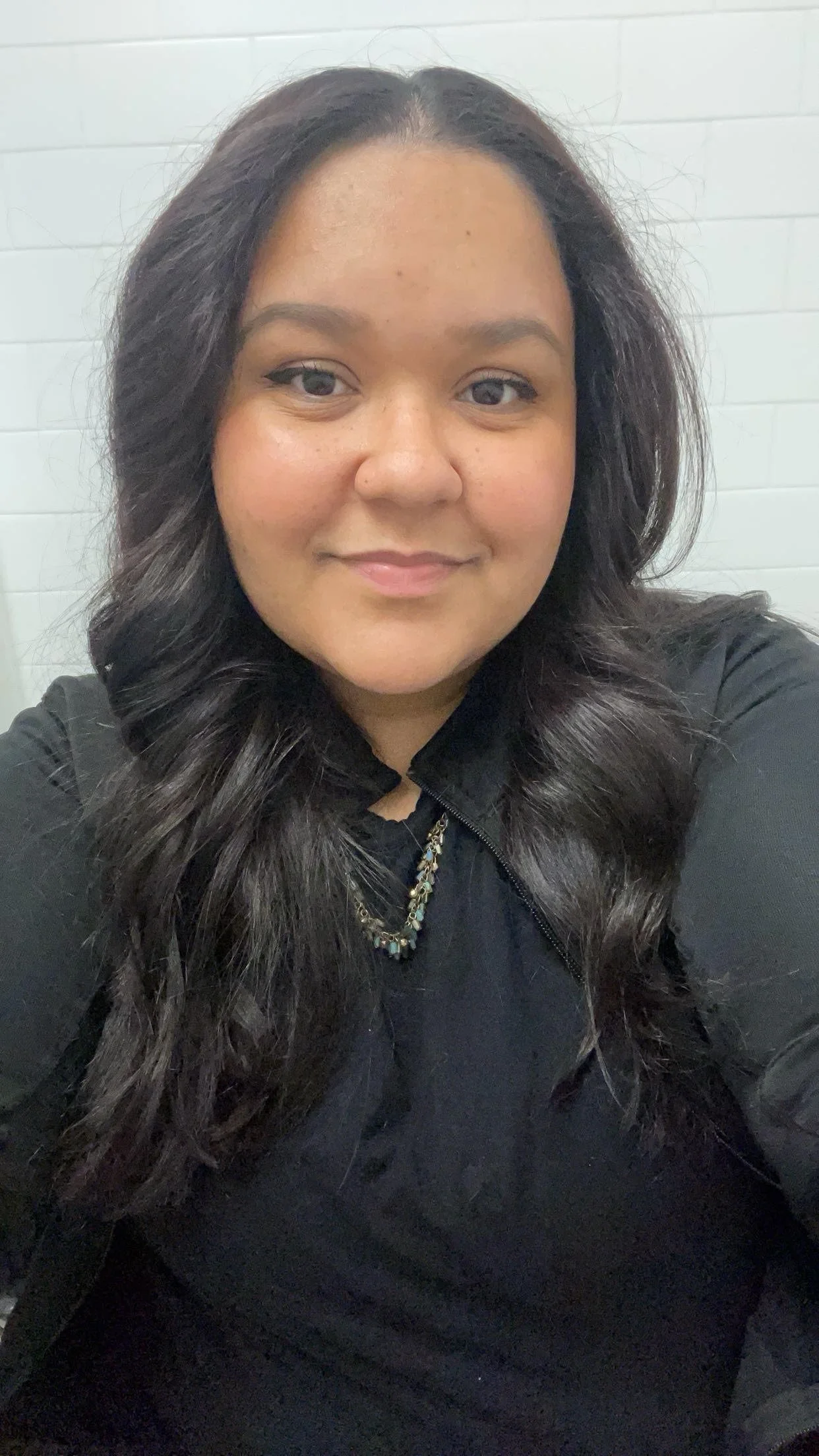  Skylynn  Rosario, a woman with long dark hair smiling at the camera, wearing a black top and a necklace, against a background of white brick walls.