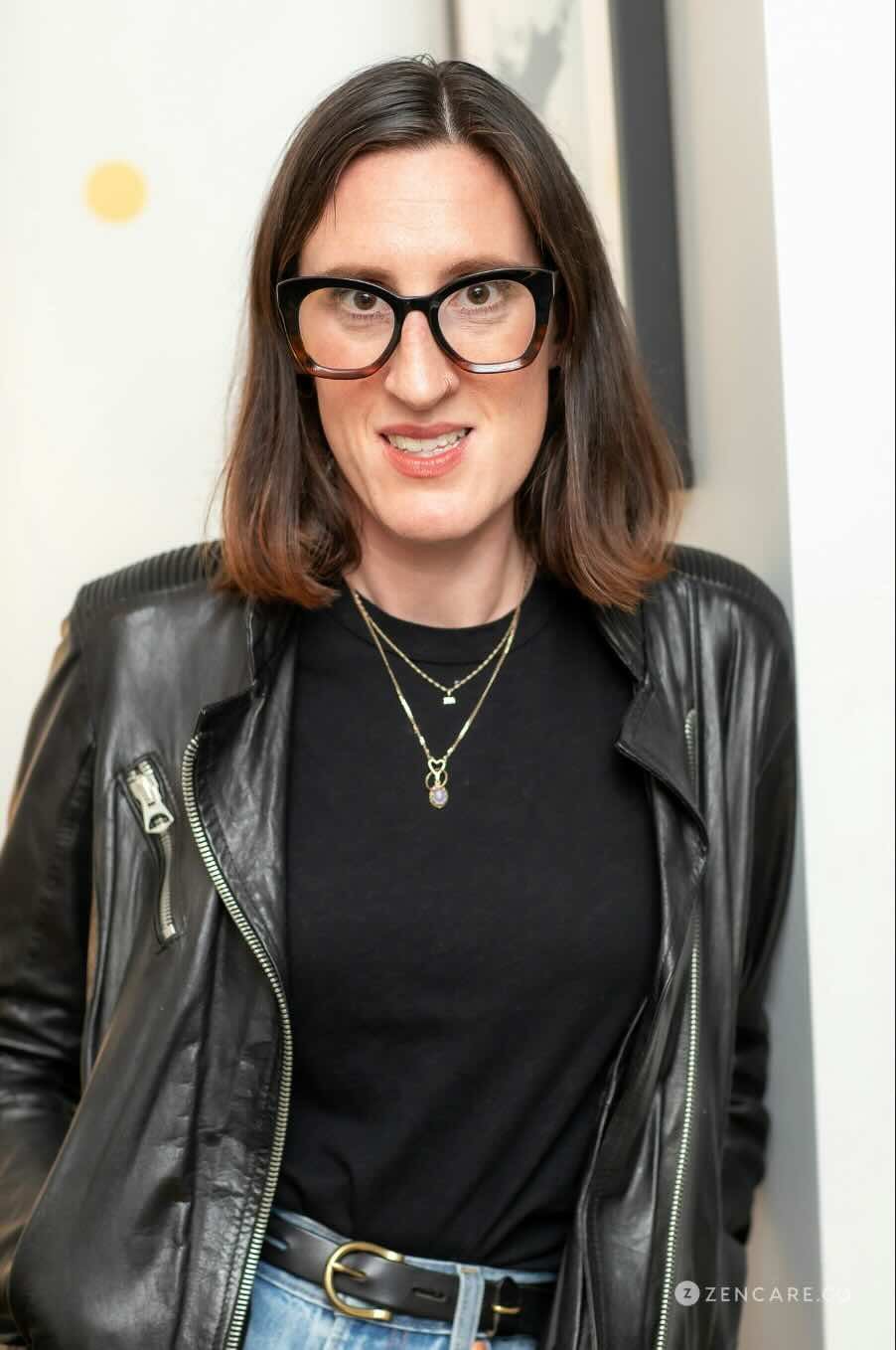 Ashley Leeds, a woman with shoulder-length brown hair, wearing large black glasses, a black leather jacket, and layered necklaces, smiling and standing in front of a white wall.