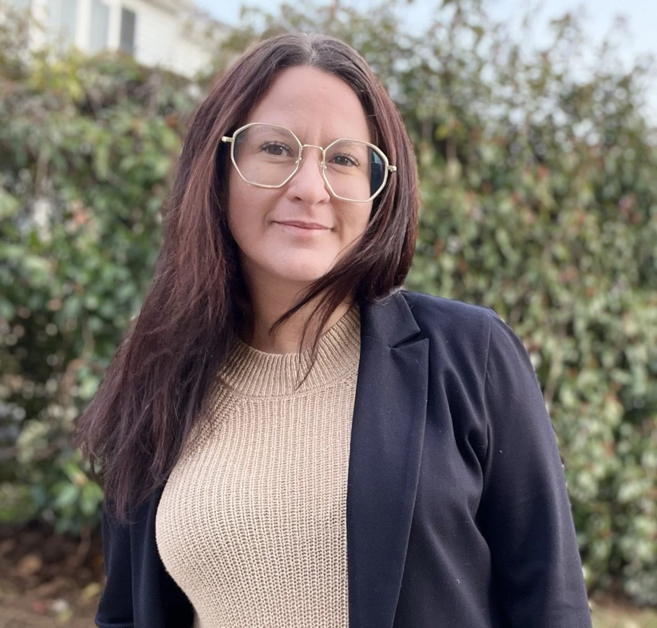 Brittany Gonzalez, a woman with dark hair and glasses, wearing a beige sweater and black blazer and standing outdoors in front of greenery.