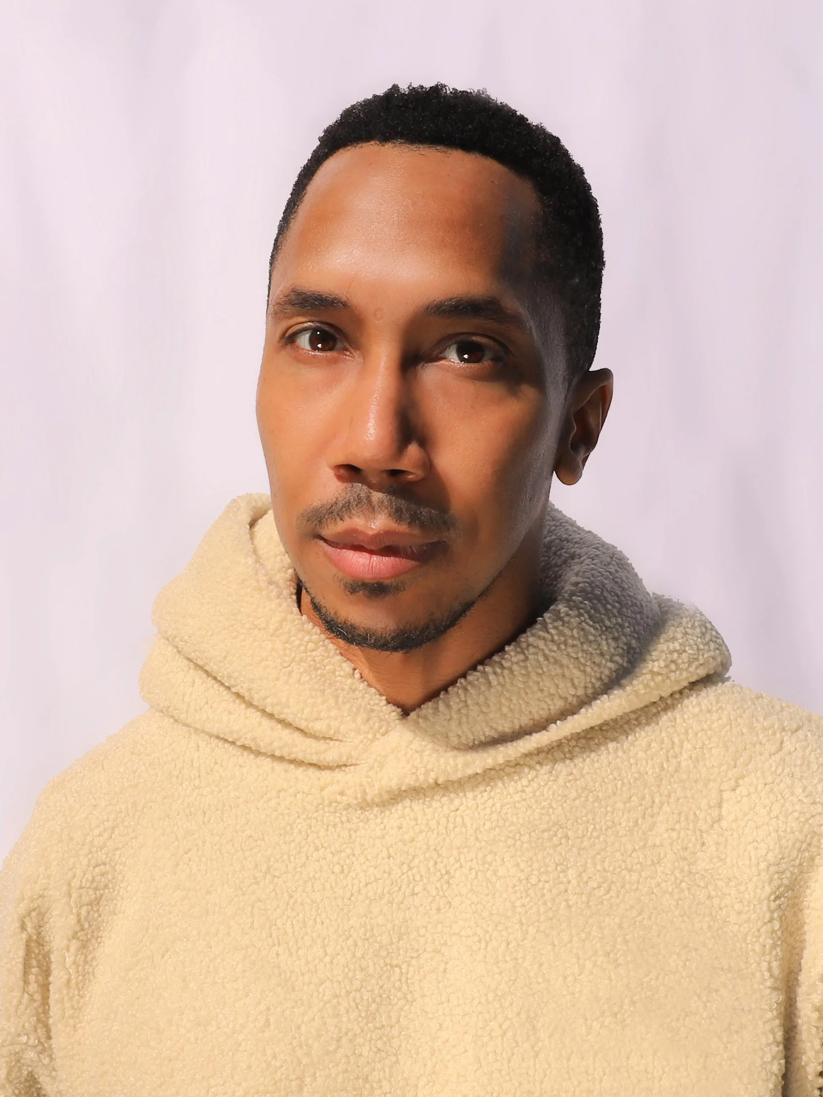 Aaron Arthur-Gray, a young man with short curly black hair and brown eyes wearing a beige fleece hoodie, standing against a white background.
