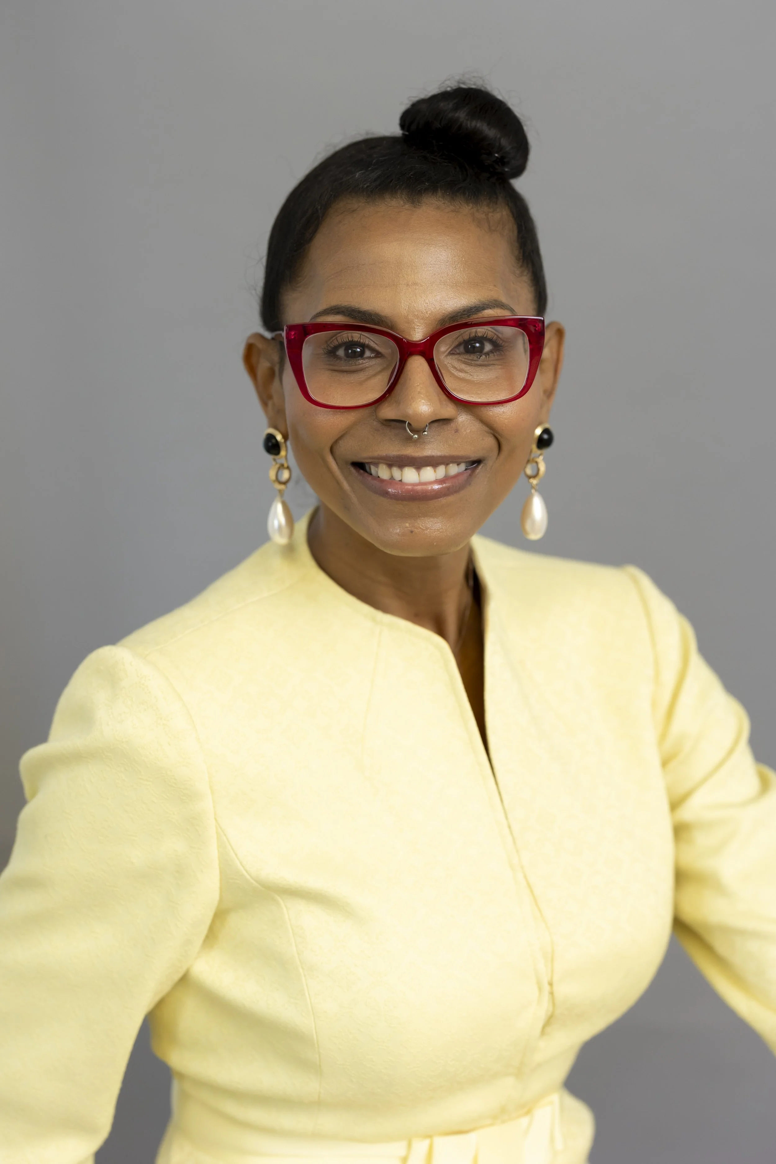 Abigail Skinner, a smiling woman with dark hair in a bun, red glasses, earrings, and a yellow blazer standing against a gray background.