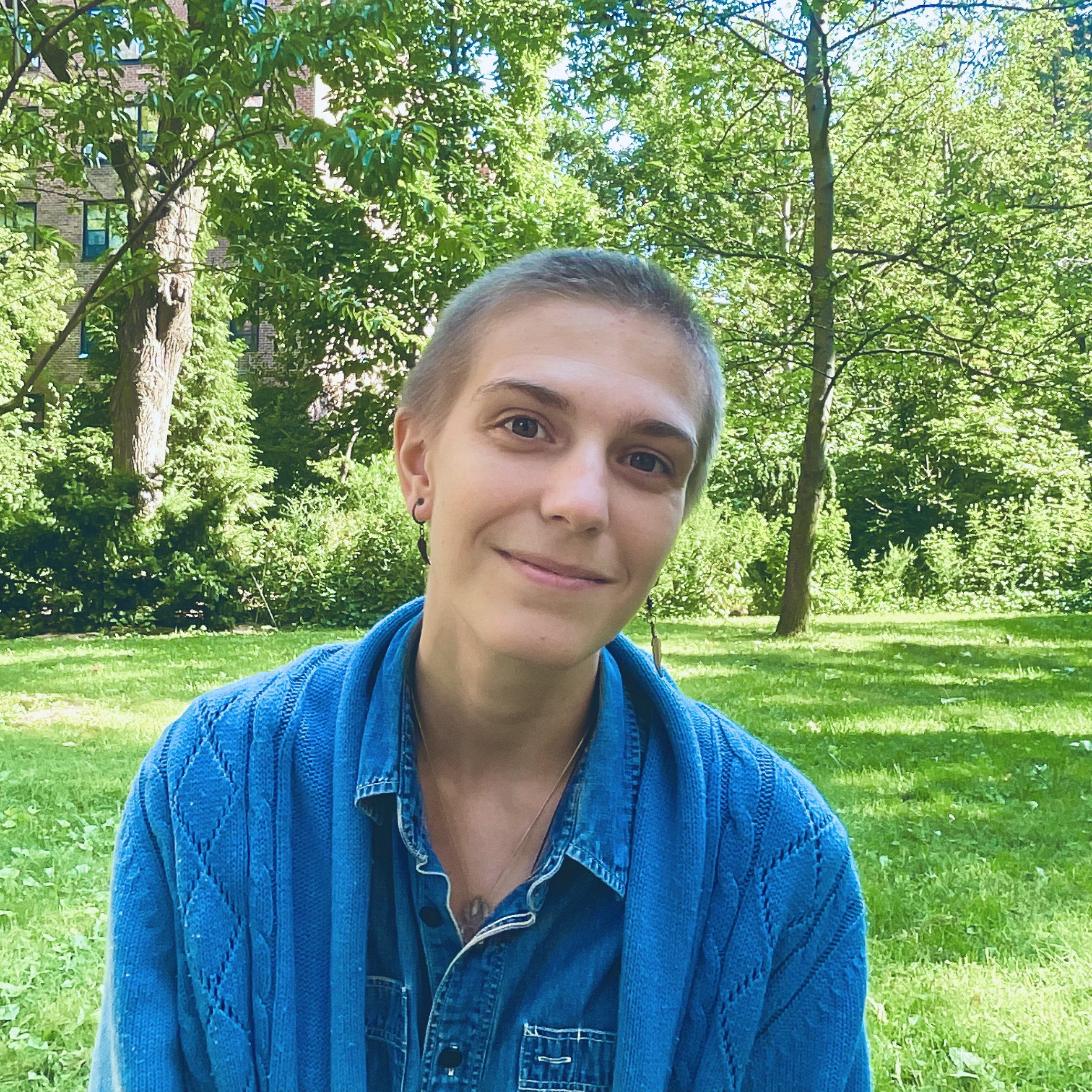 A person with short hair and earrings smiling outdoors, wearing a blue denim jacket, in a park with green grass and trees.