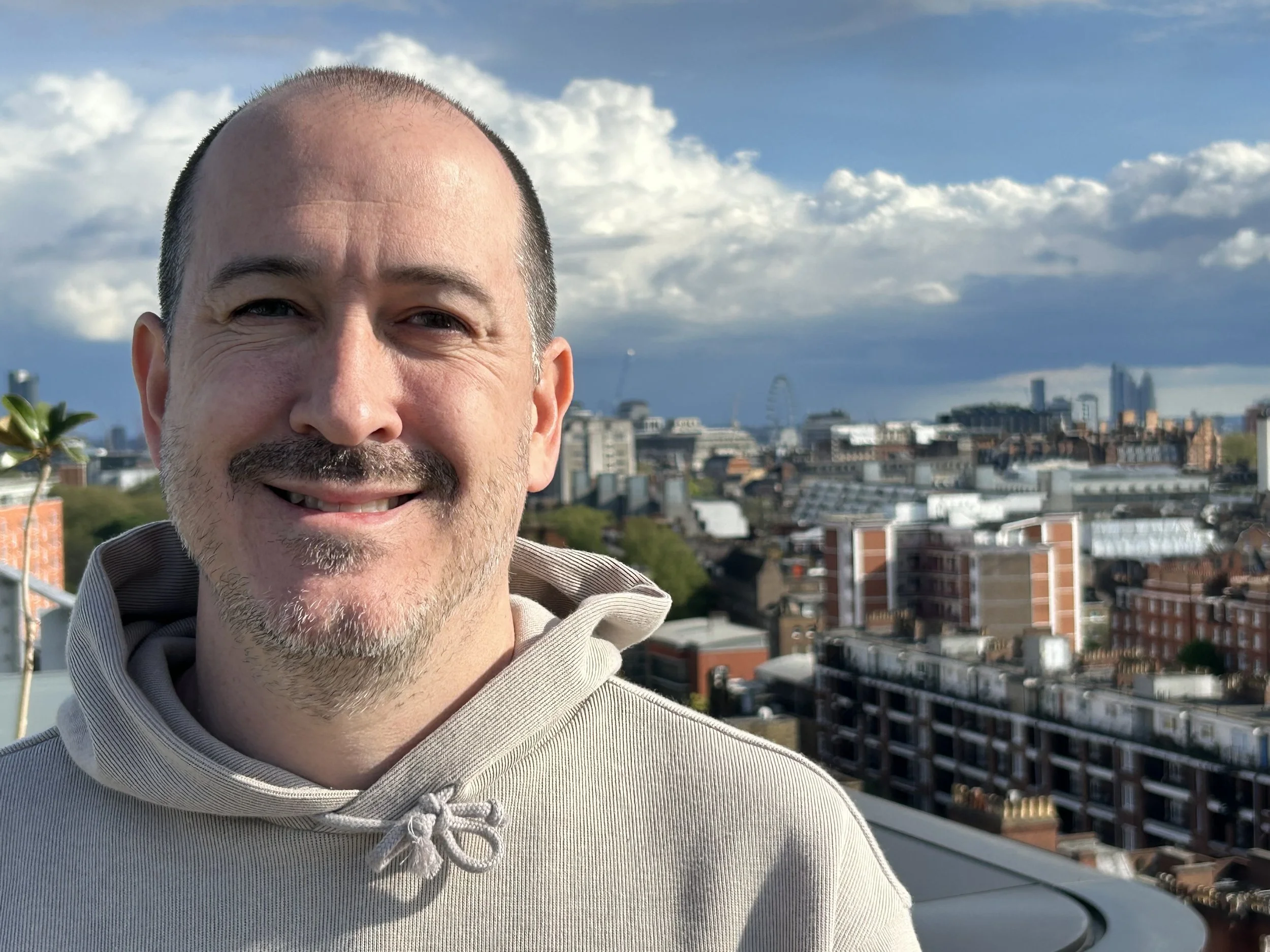 Kevin Skinner-Spain, a smiling man with a beard and short hair, wearing a beige hoodie, standing on a rooftop with a city skyline in the background.