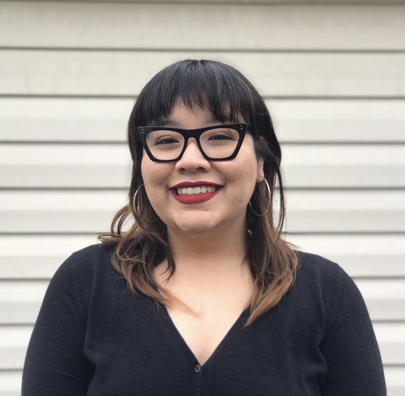 Stephanie Del Cid, a woman with dark hair and light brown highlights, wearing glasses, hoop earrings, red lipstick, and a black top, smiling in front of a beige outdoor wall.