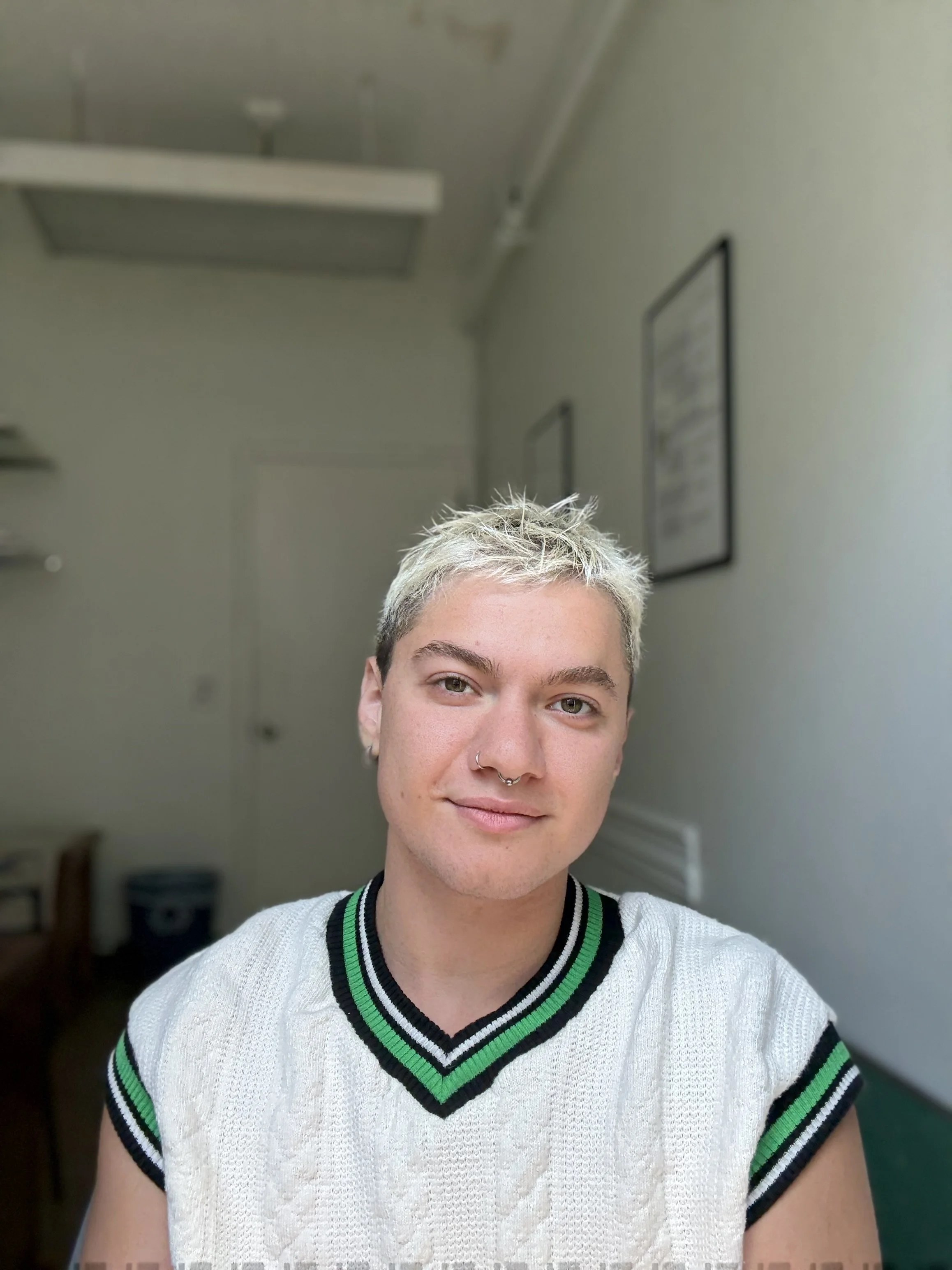 River Weisz, a young person with short, platinum blonde hair and a septum ring, wearing a white sleeveless sweater with green and black stripes.