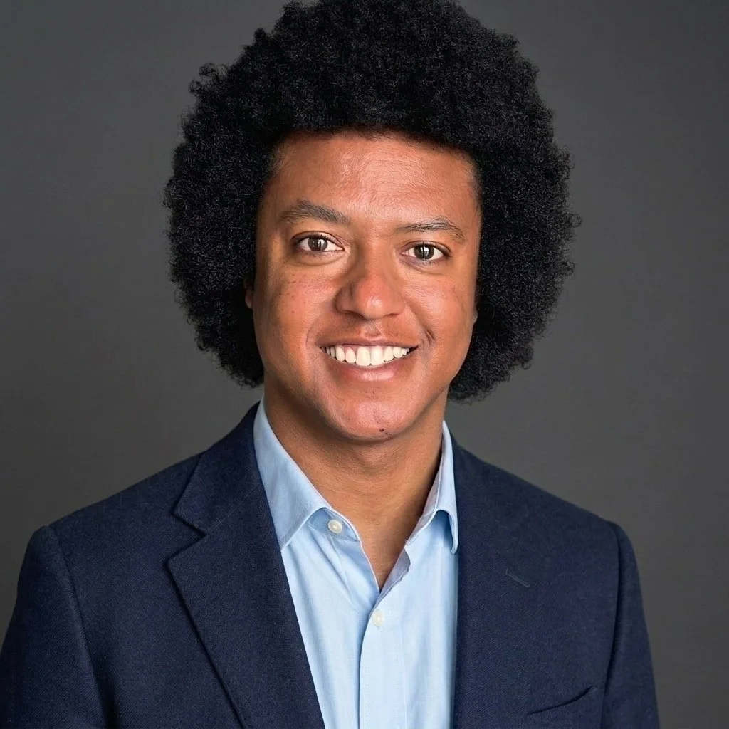 Headshot of Aaron Skinner-Spain, a smiling young man wearing a purple shirt and black tie against a dark background.