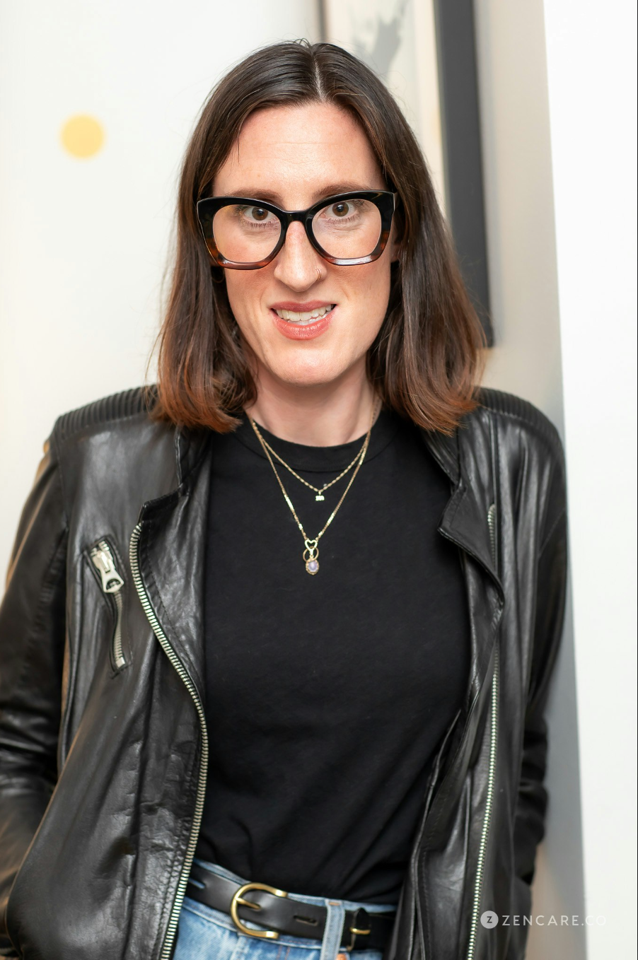 A woman with shoulder-length brown hair wearing large black glasses, a black leather jacket, a black t-shirt, layered necklaces, and blue jeans, standing indoors near a white wall.