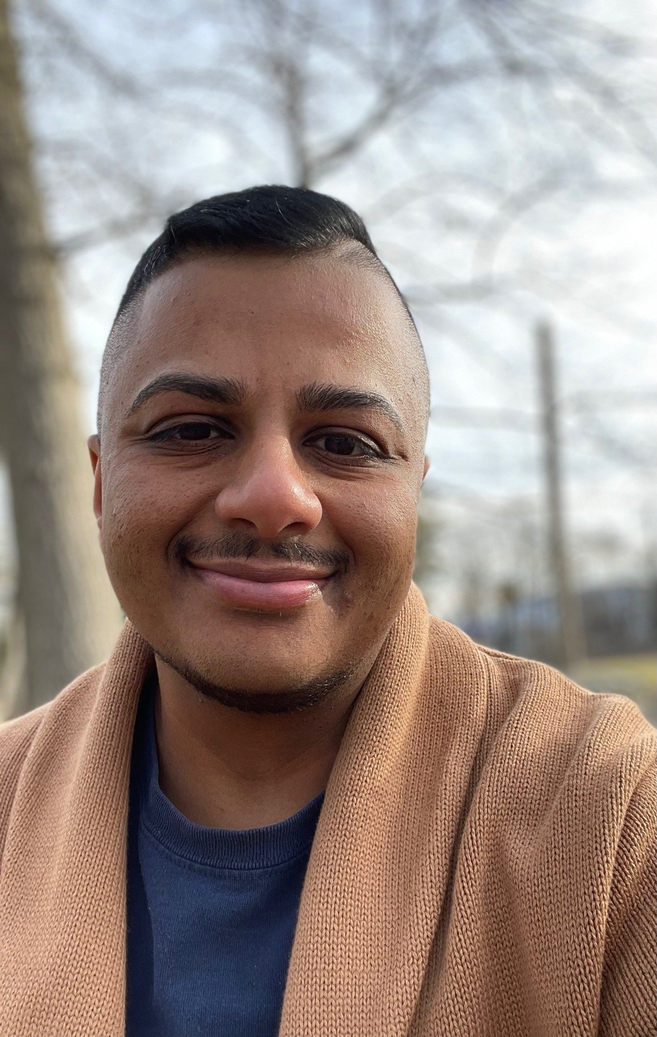 David Chary, a smiling man with short black hair, tan skin, and a goatee, wearing a tan jacket over a dark blue shirt, outdoors with sky in the background.
