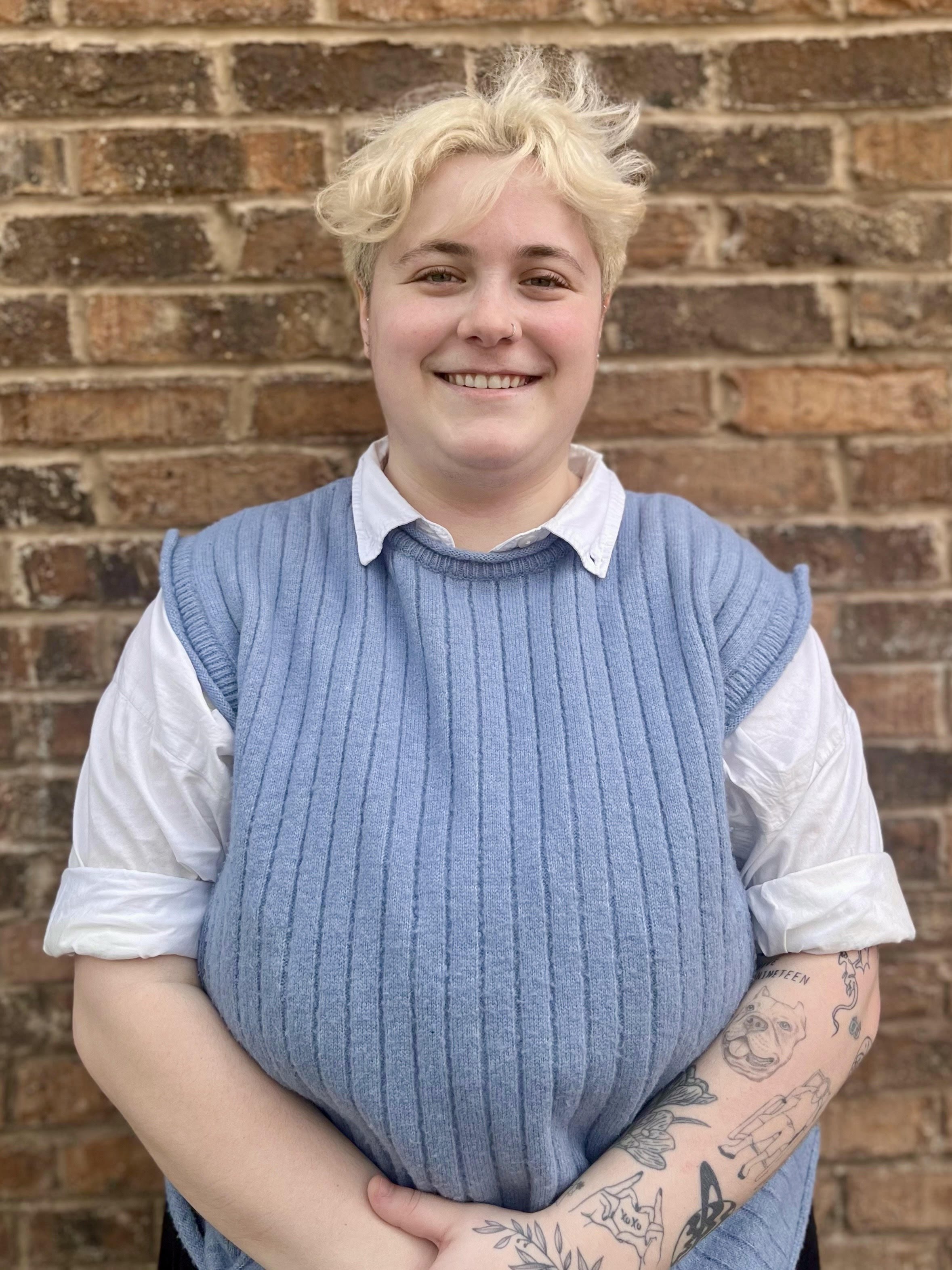 Sophie Kaplan, a person with short, curly, blonde hair standing in front of a brick wall. They are smiling and wearing a white shirt with rolled-up sleeves under a blue knitted vest. They have a nose ring and tattoos on one forearm.