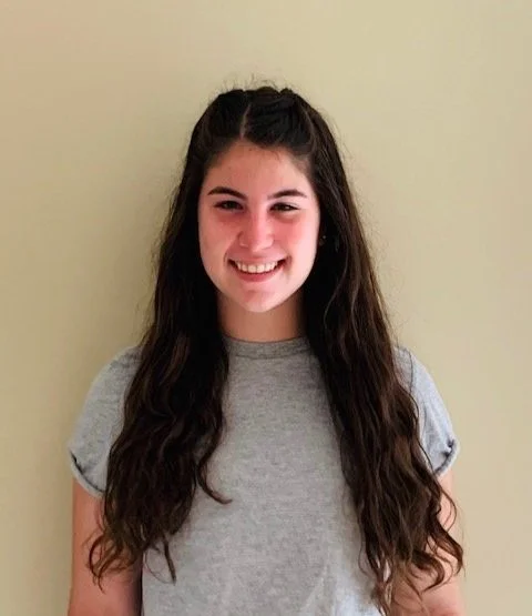 Jenna Kupferschmid, a young woman with long dark hair smiling, wearing a gray t-shirt, standing against a light-colored wall.