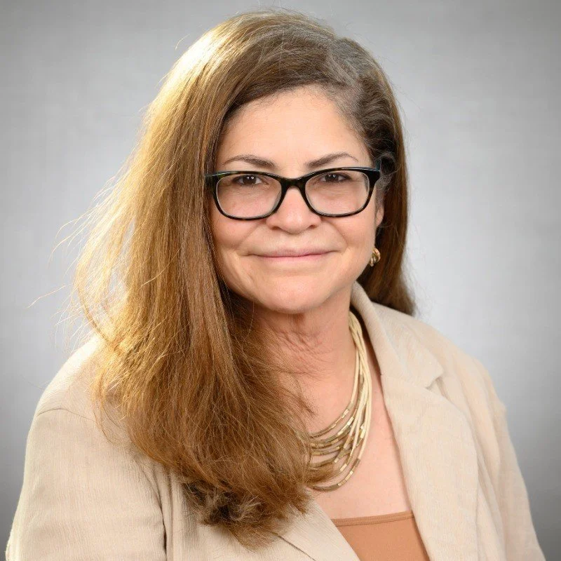 A woman with light brown, wavy hair, wearing glasses, and a beige blazer, smiling at the camera with a gray background.