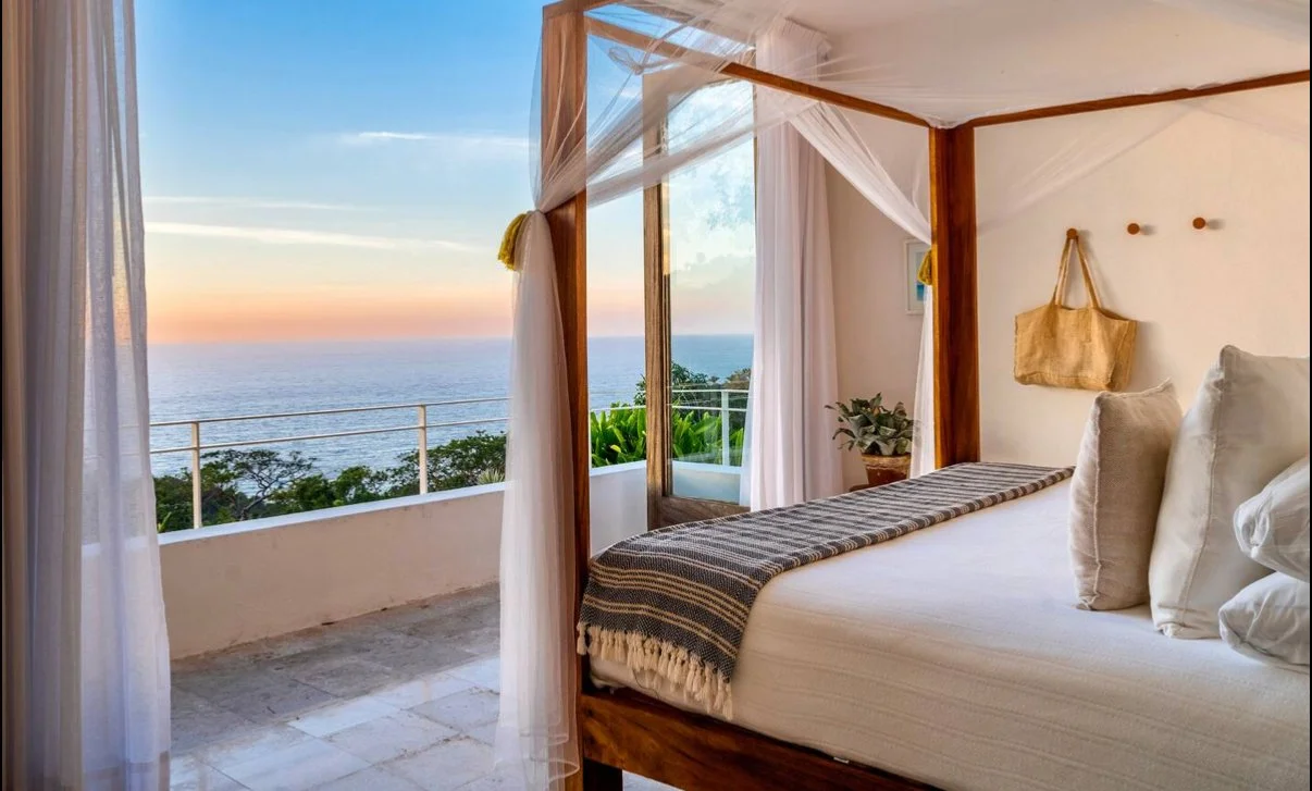 A bedroom with a four-poster bed and netting, overlooking an ocean view at sunset.